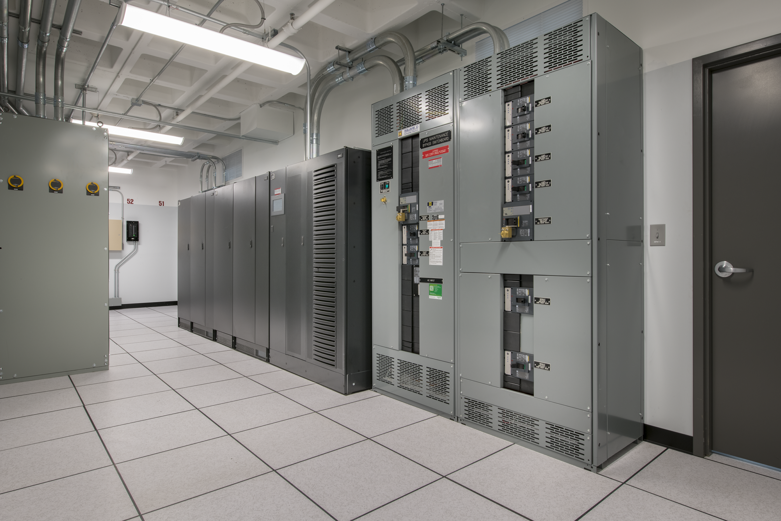A clean server or electrical room with gray equipment racks, control panels, and large cabinets on tiled flooring. Overhead pipes and fluorescent lights are visible. A closed gray door is on the right wall.
