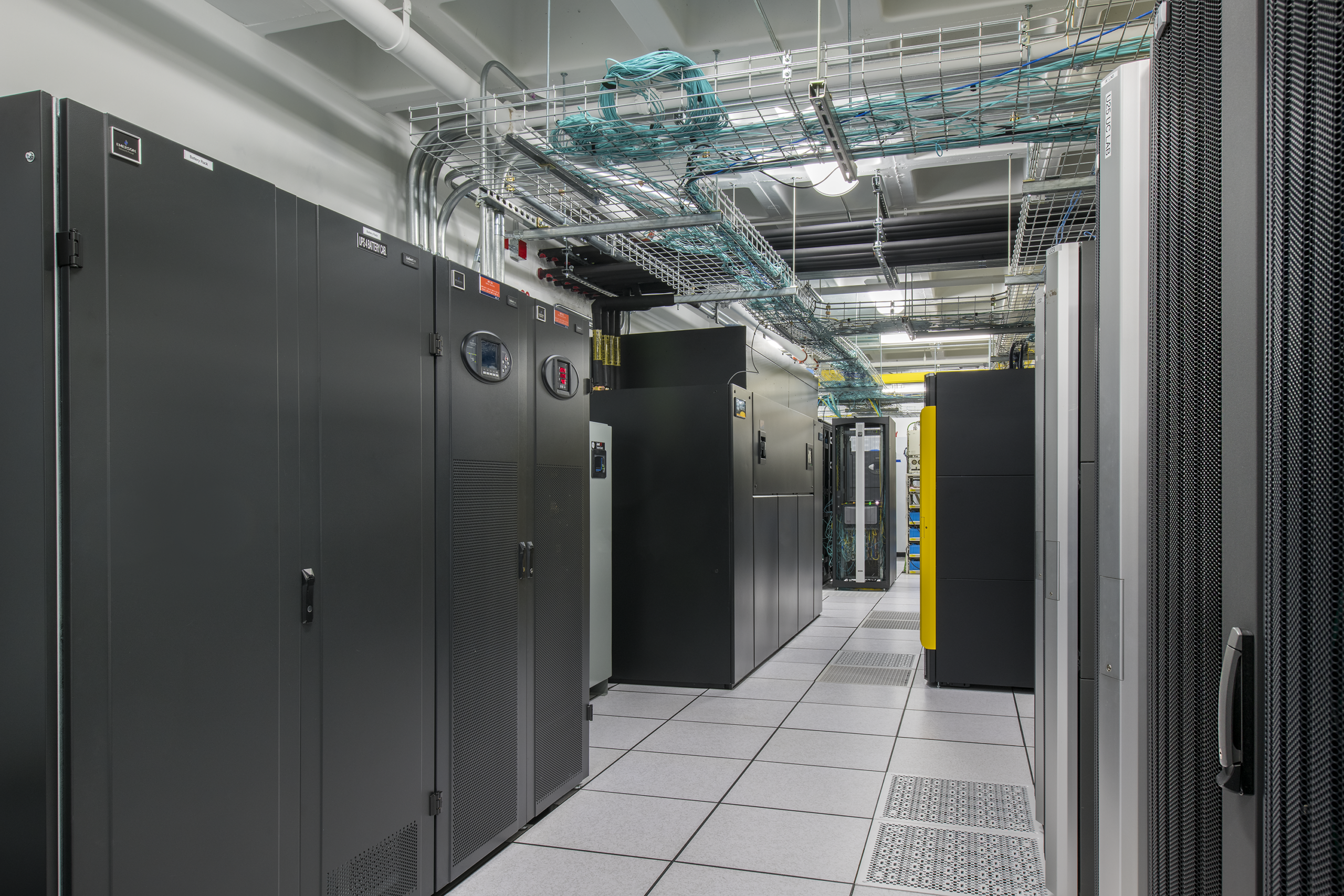 A modern data center with rows of black server racks, cable trays on the ceiling, and a tiled floor. The room is brightly lit, with visible network cables and equipment neatly organized.