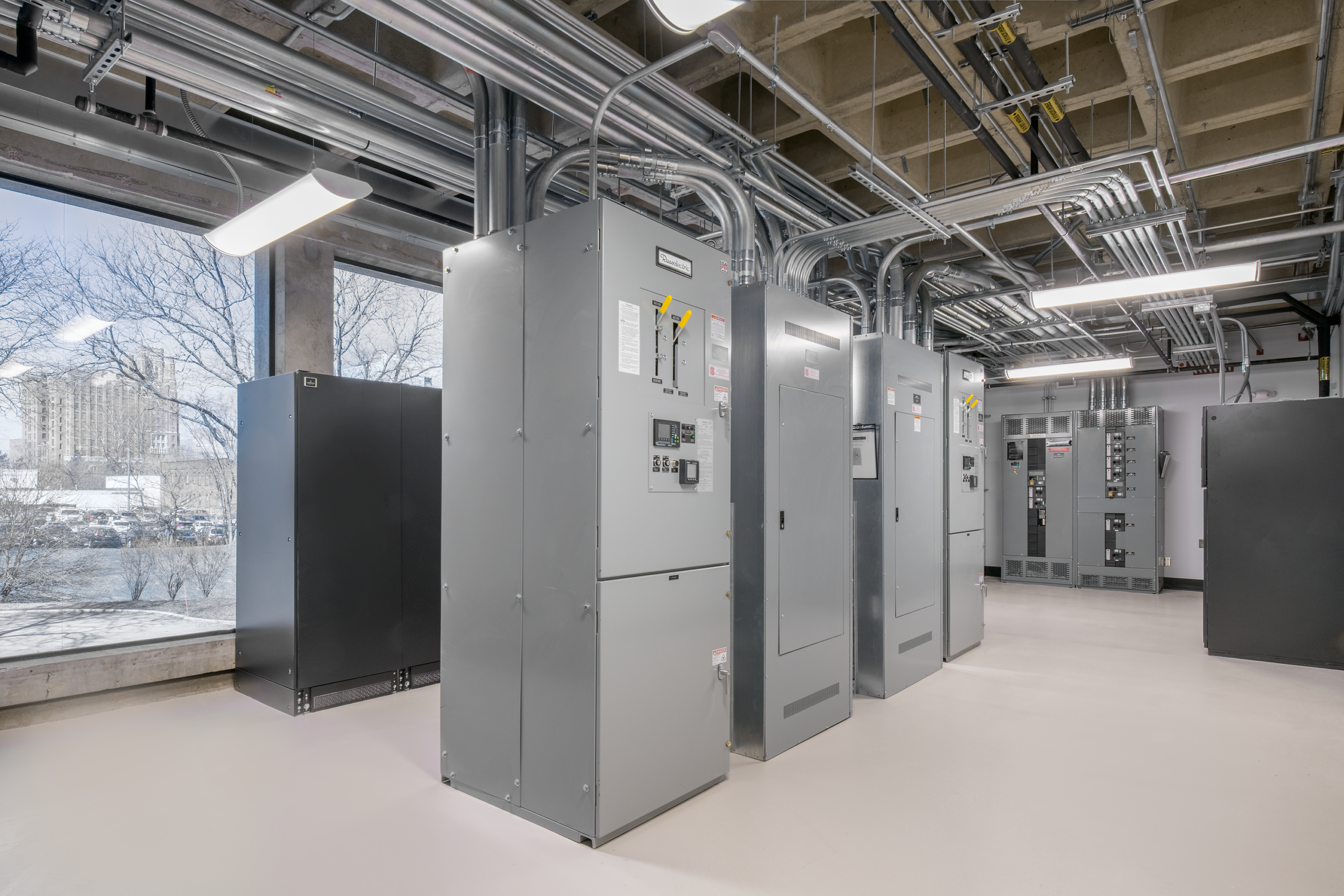 Rows of large gray electrical control panels and circuit breaker boxes line a clean, bright utility room with exposed pipes and large windows showing trees and buildings outside.