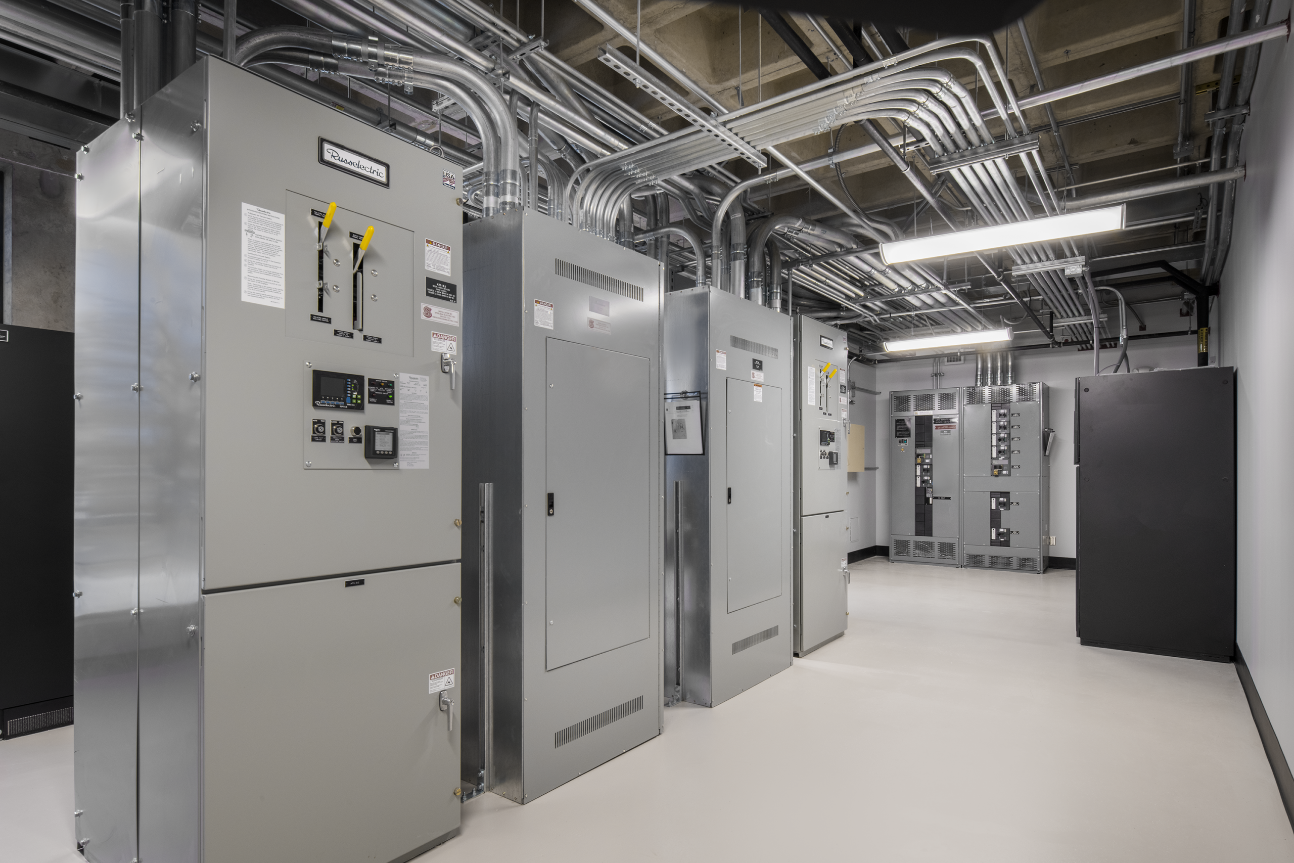 A room with large gray electrical panels, circuit breakers, and numerous overhead metal conduit pipes, all clean and neatly organized under bright ceiling lights.