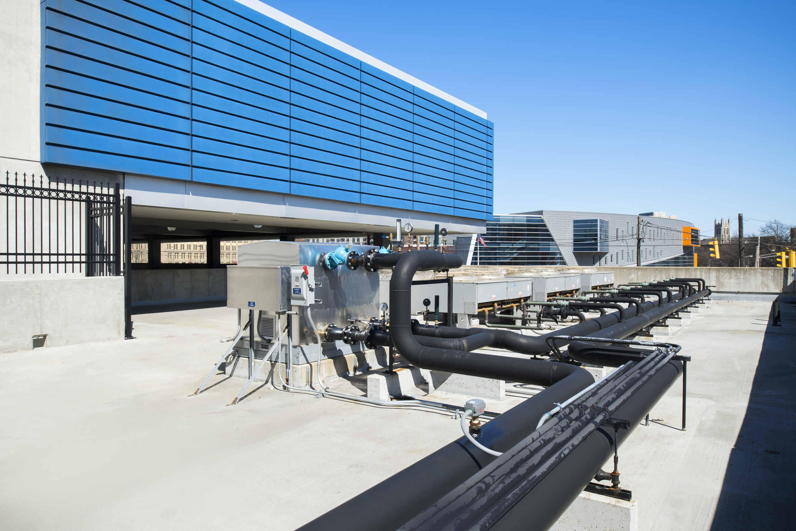 A rooftop with black pipes, electrical boxes, and industrial equipment beneath a blue building facade on a clear sunny day.