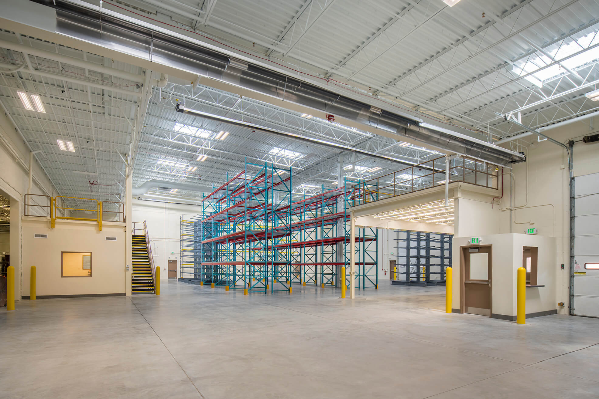 A spacious, clean warehouse interior with empty blue and red metal storage racks, high ceilings, bright lighting, concrete floors, and a small office area to the left with stairs and yellow safety railings.