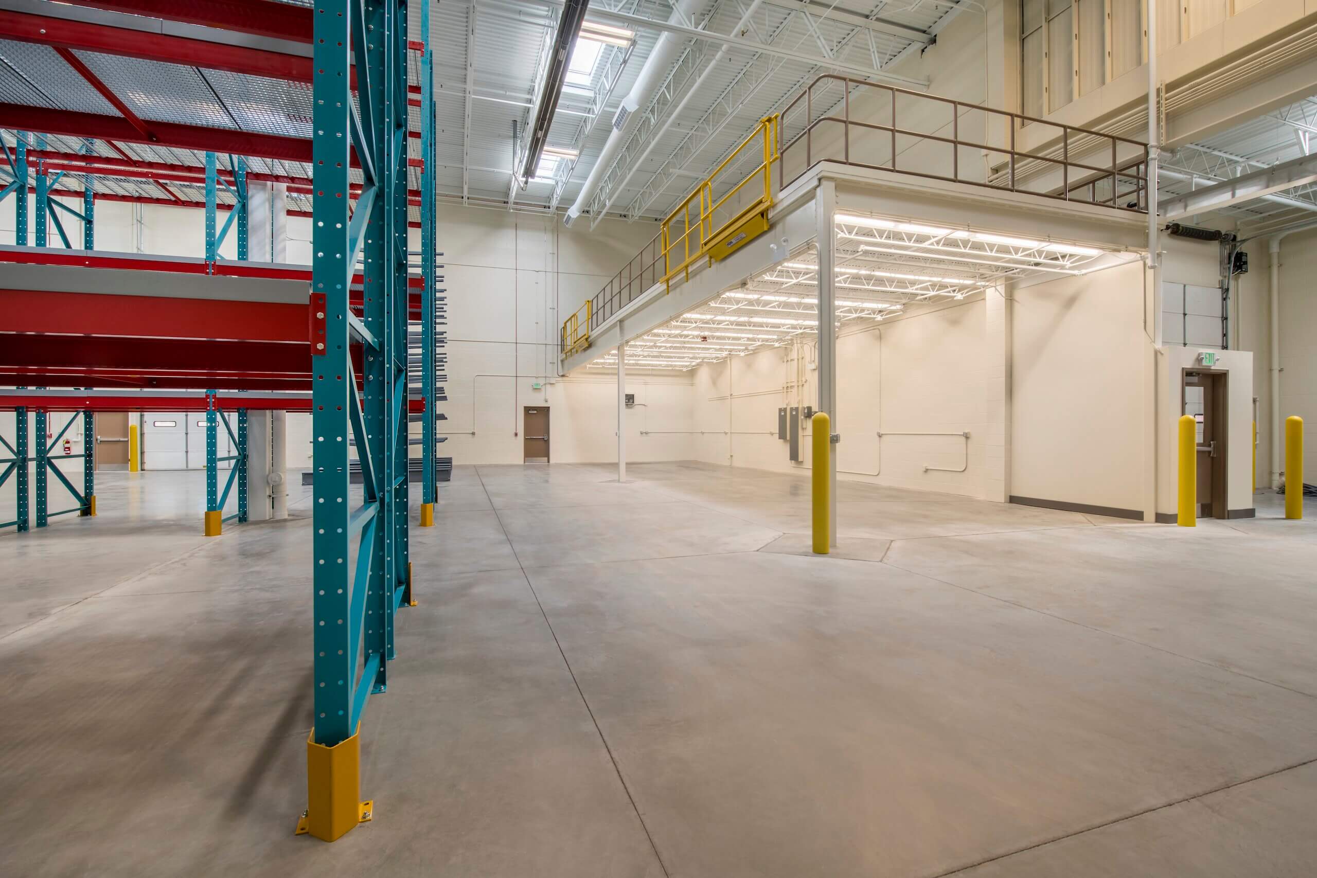 Empty warehouse interior with high ceilings, bright lighting, concrete floors, metal shelving units on the left, a yellow-railed mezzanine on the right, and yellow safety bollards near the entrance.