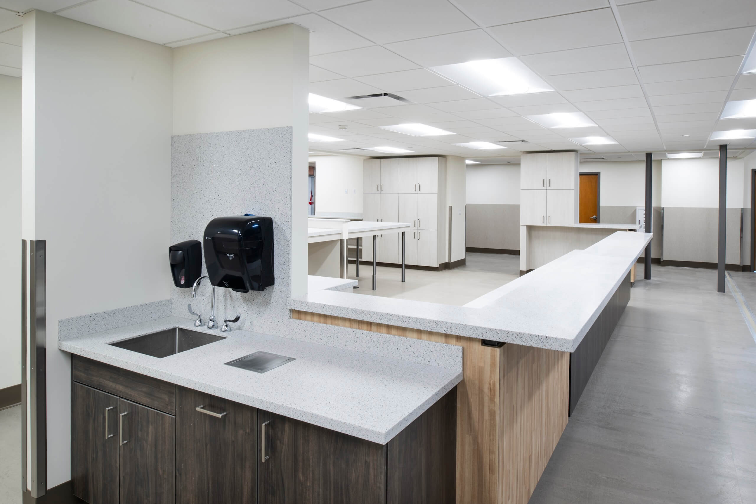 Modern, clean medical or laboratory workspace with sinks, soap dispensers, white countertops, and overhead lighting. Cabinets and workstations are visible throughout the room.