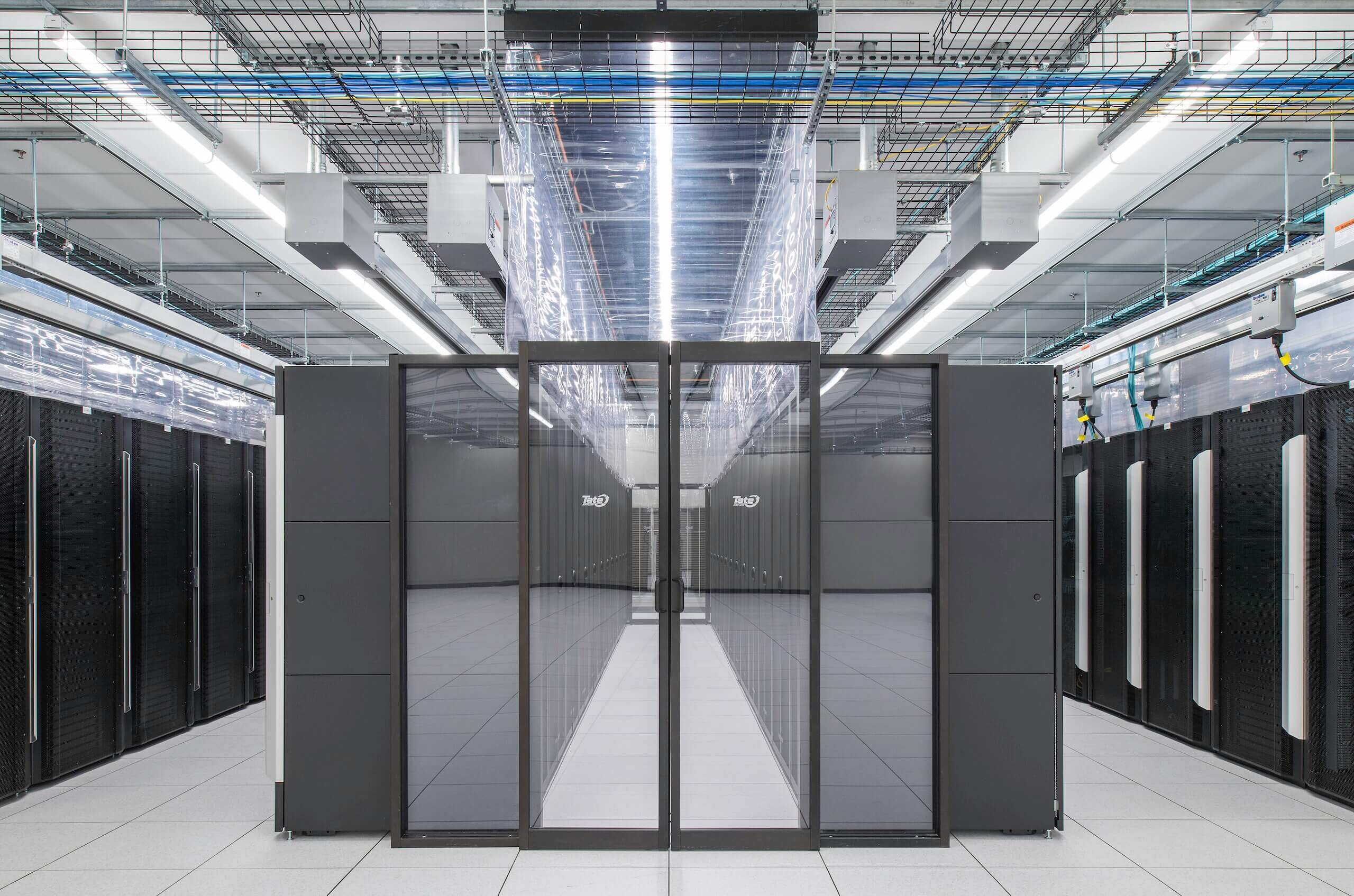 A modern data center with rows of black server racks, overhead cable trays, and bright lighting. The floor is white and reflective, and glass doors are visible in the center aisle.