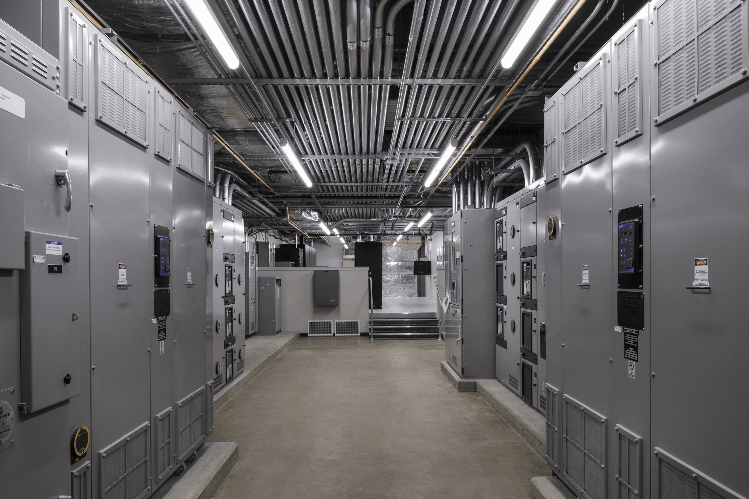 A modern industrial control room with gray electrical panels lining both sides, exposed pipes and wires on the ceiling, bright fluorescent lighting, and a clean concrete floor.