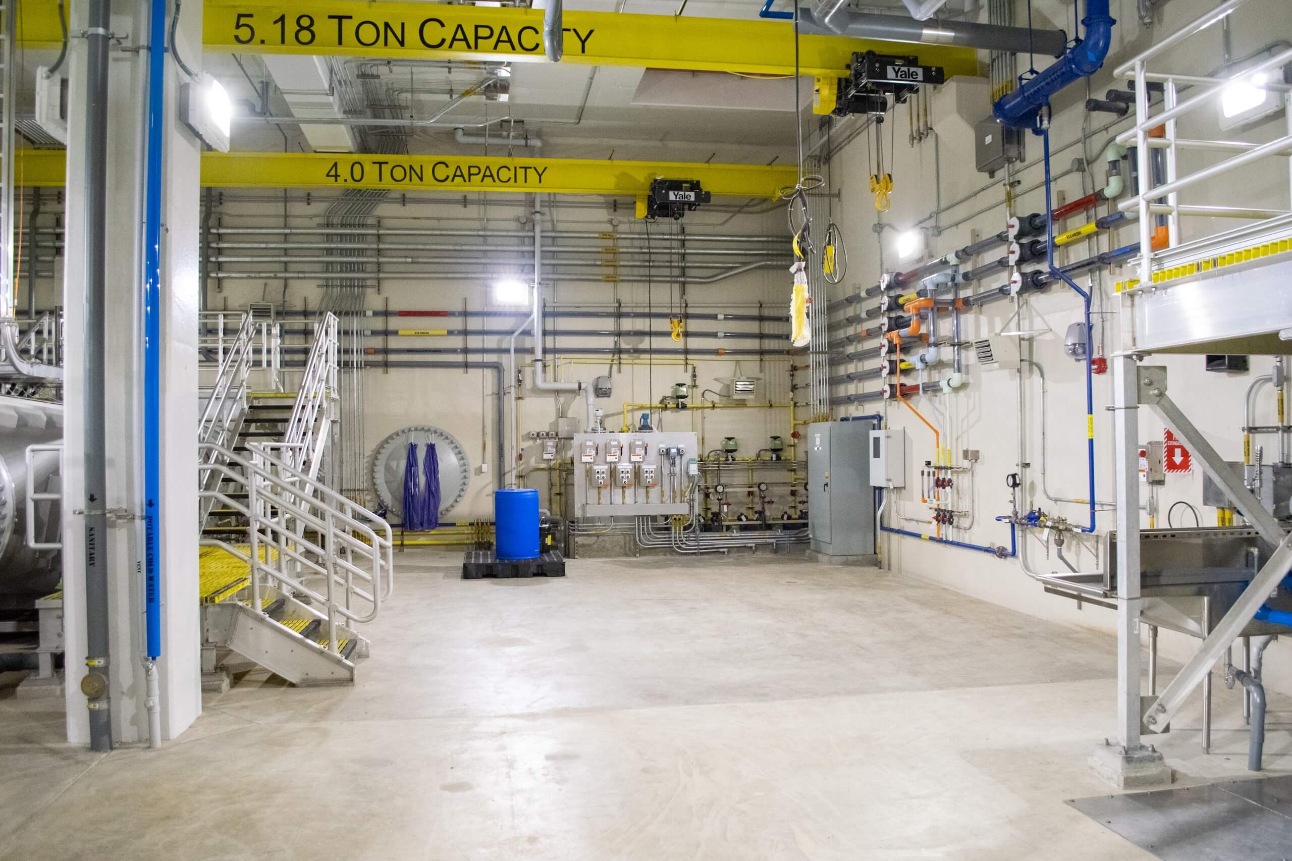 Industrial facility interior with metal stairs, machinery, pipes on the walls, and yellow overhead cranes labeled 5.18 TON CAPACITY and 4.0 TON CAPACITY in a clean, well-lit space.