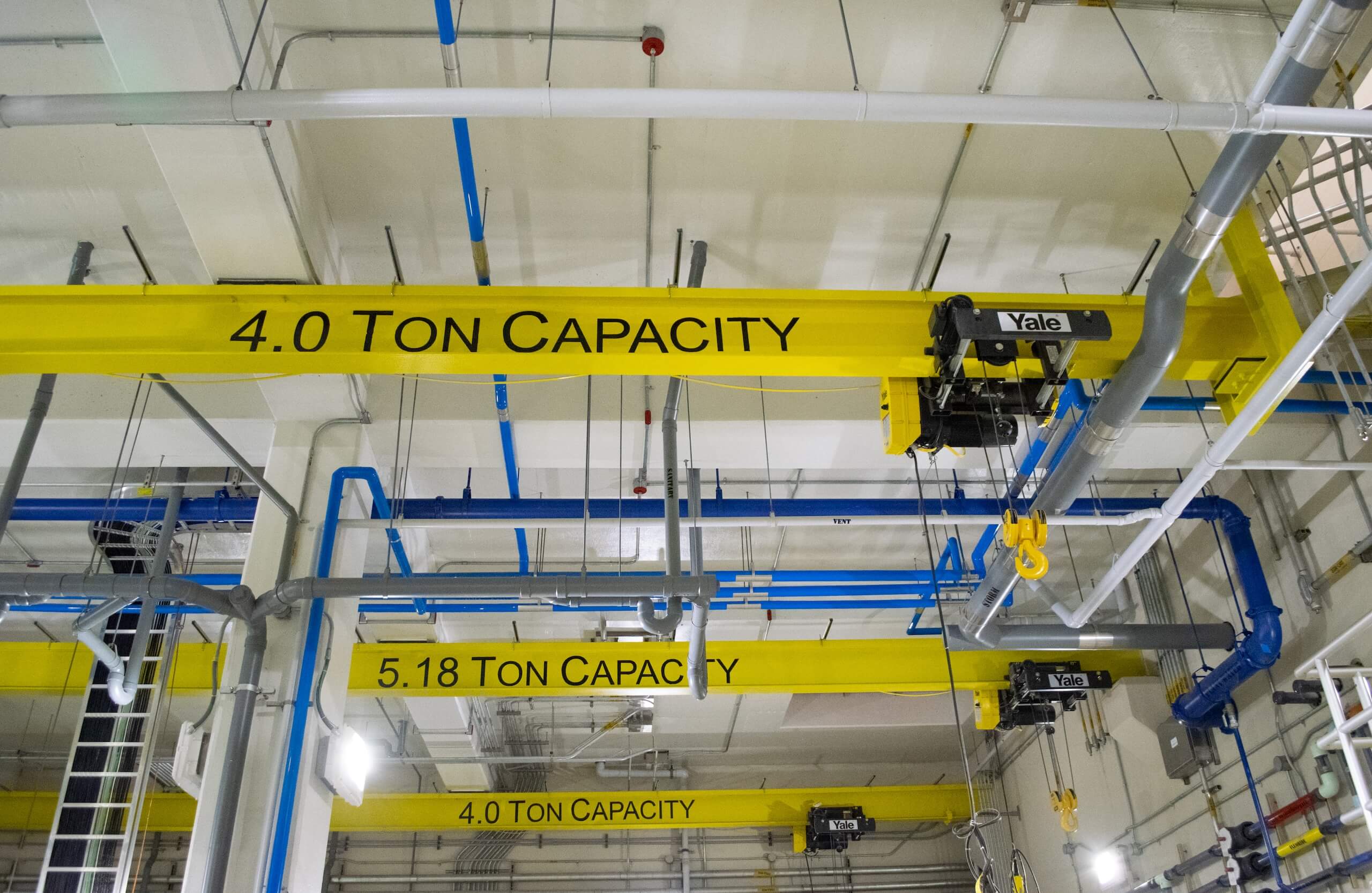 Yellow industrial cranes with 4.0 TON CAPACITY and 5.18 TON CAPACITY written on them are suspended from a factory ceiling among blue pipes and other machinery.