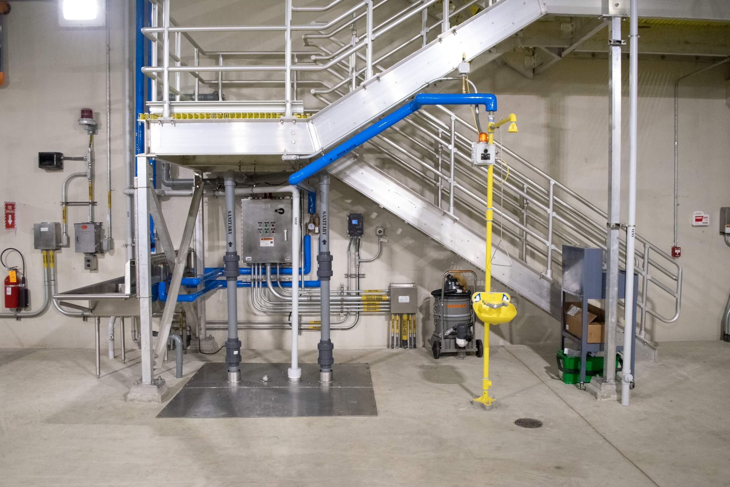 Industrial interior with metal stairs, control panels, pipes, and an emergency eye wash station with a yellow shower and basin below the stairs. Cardboard boxes and green containers are in the background.