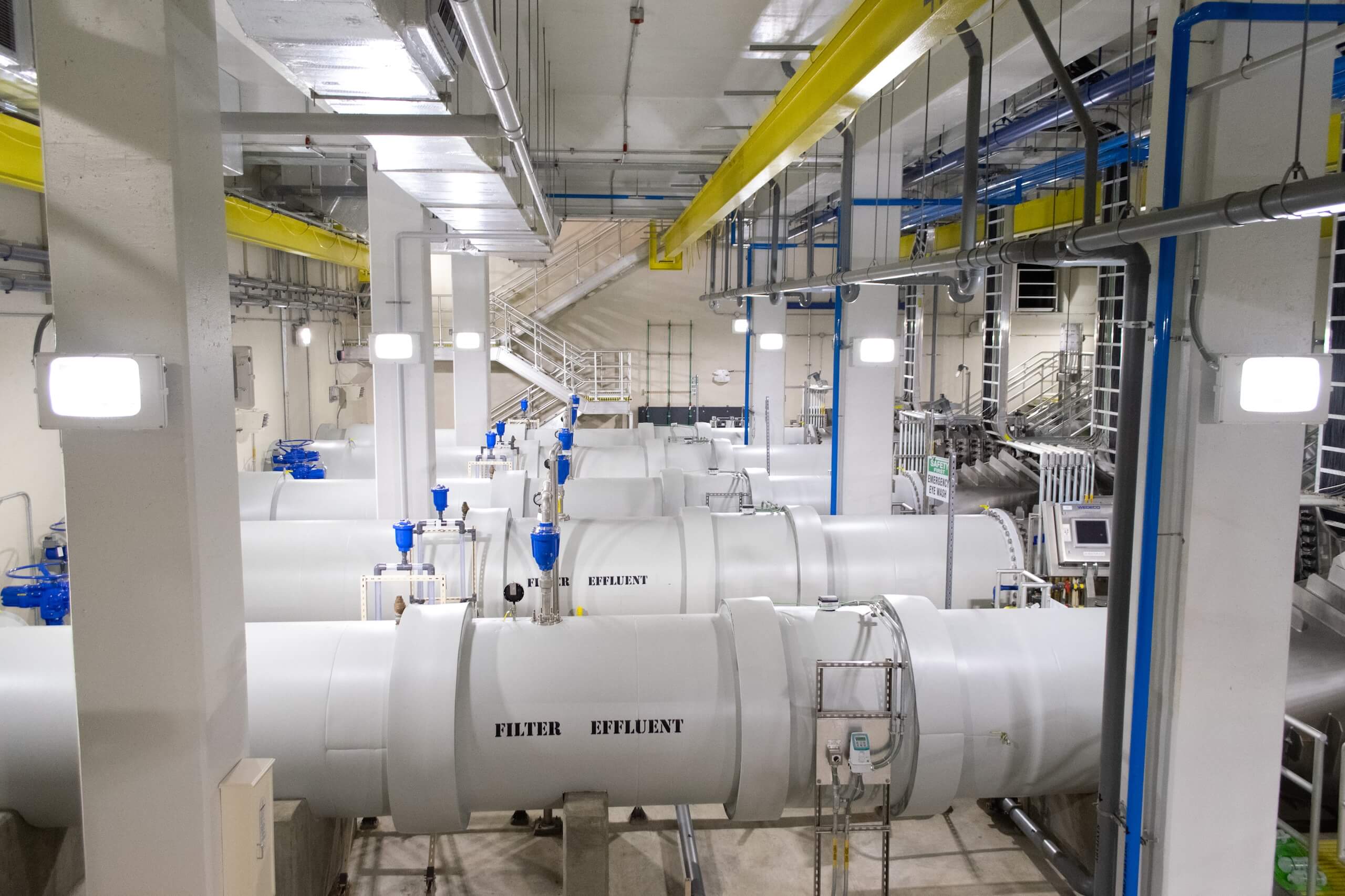 Large white industrial pipes labeled FILTER and EFFLUENT run through a brightly lit facility with blue valves, yellow ceiling beams, and various control panels and stairs visible in the background.