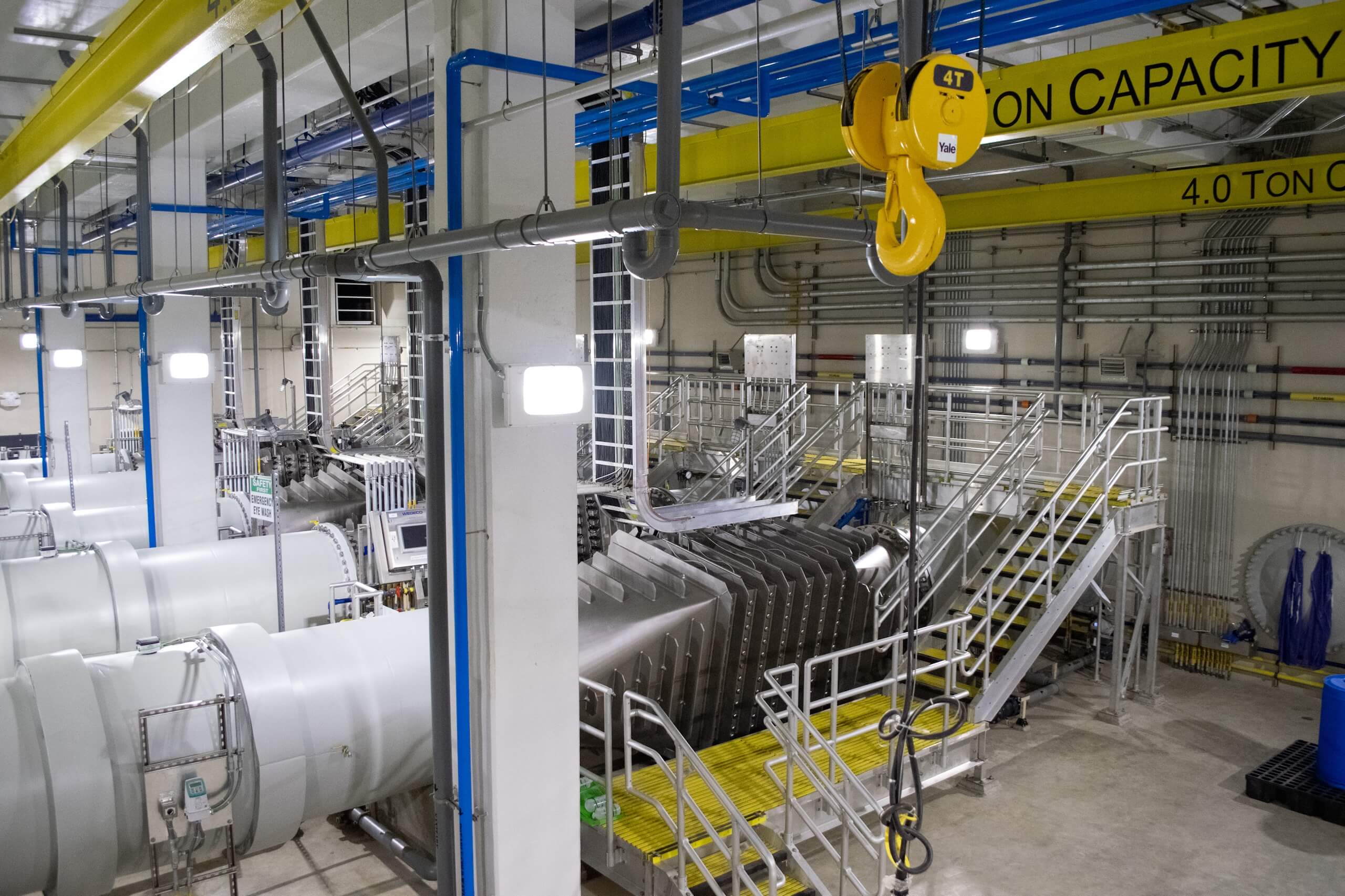 An industrial facility with metal stairs, platforms, pipes, ducts, and a yellow overhead crane labeled 4.0 ton capacity in a brightly lit setting.
