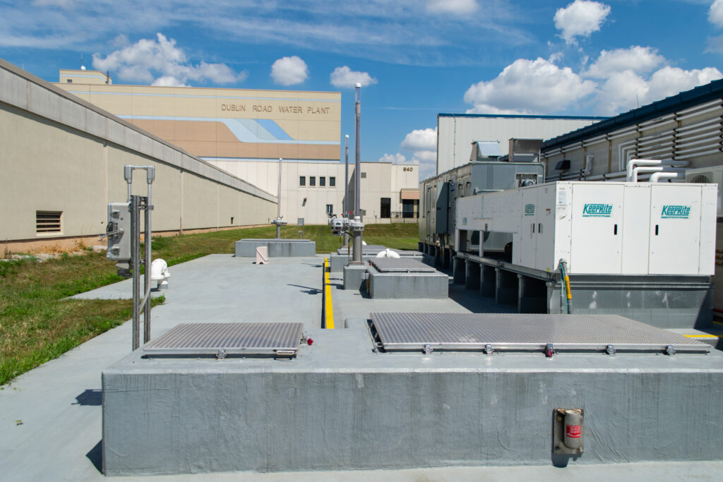 Outdoor view of the Dublin Road Water Plant showing industrial equipment, large backup generators, metal grates, and utility structures under a blue sky with scattered clouds.