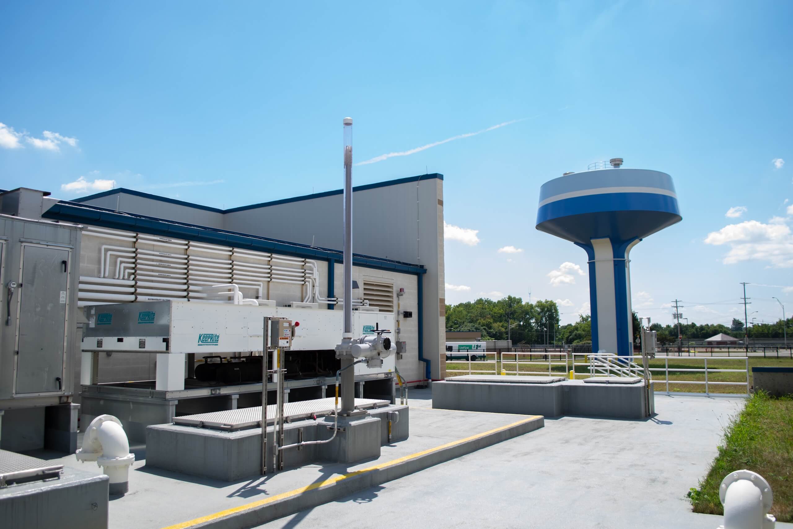 A modern water treatment facility with large equipment in the foreground and a blue-and-white water tower in the background under a clear, sunny sky.