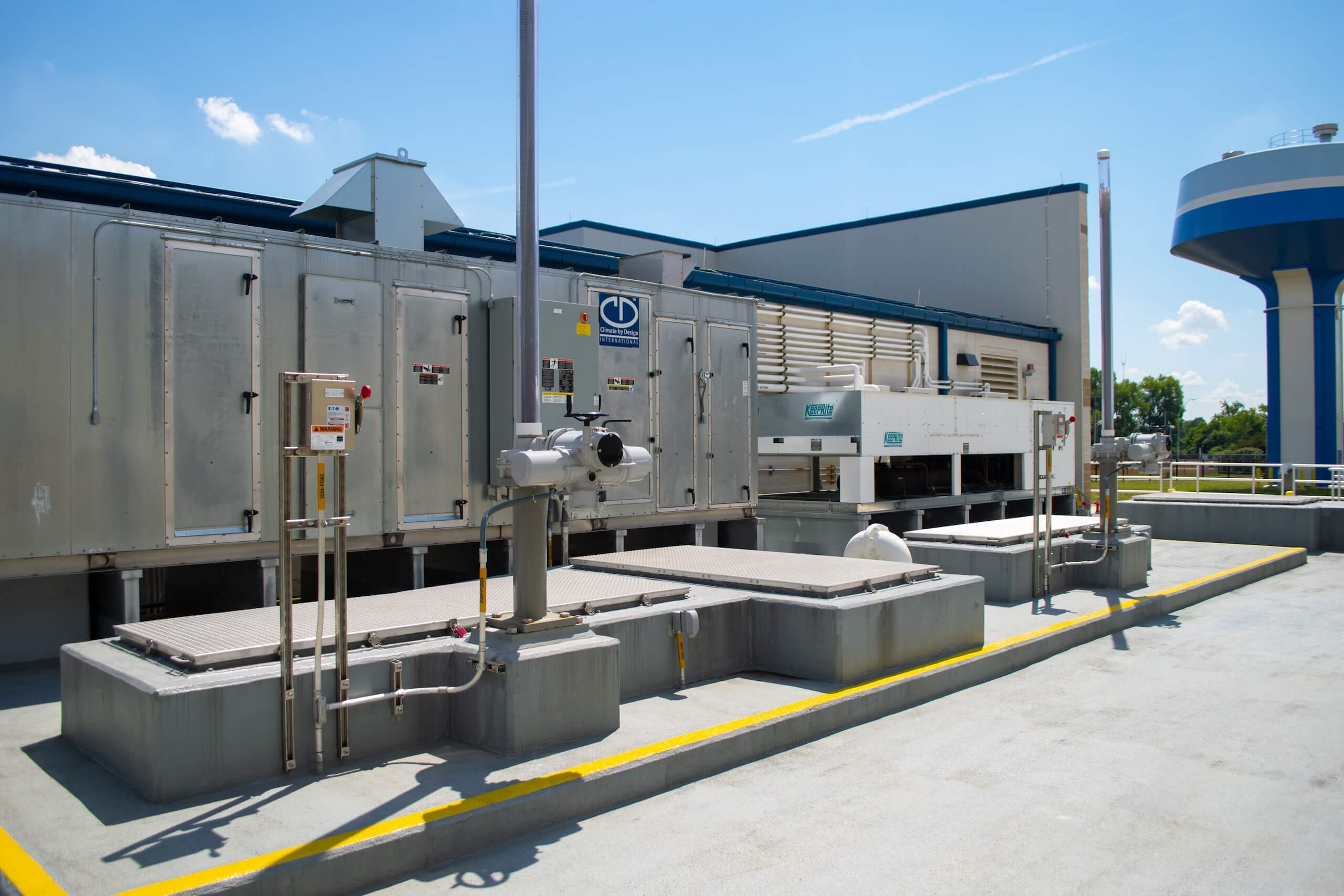 Large industrial HVAC units and equipment sit on a rooftop, surrounded by railings and pipes, under a bright blue sky with a water tower visible in the background.