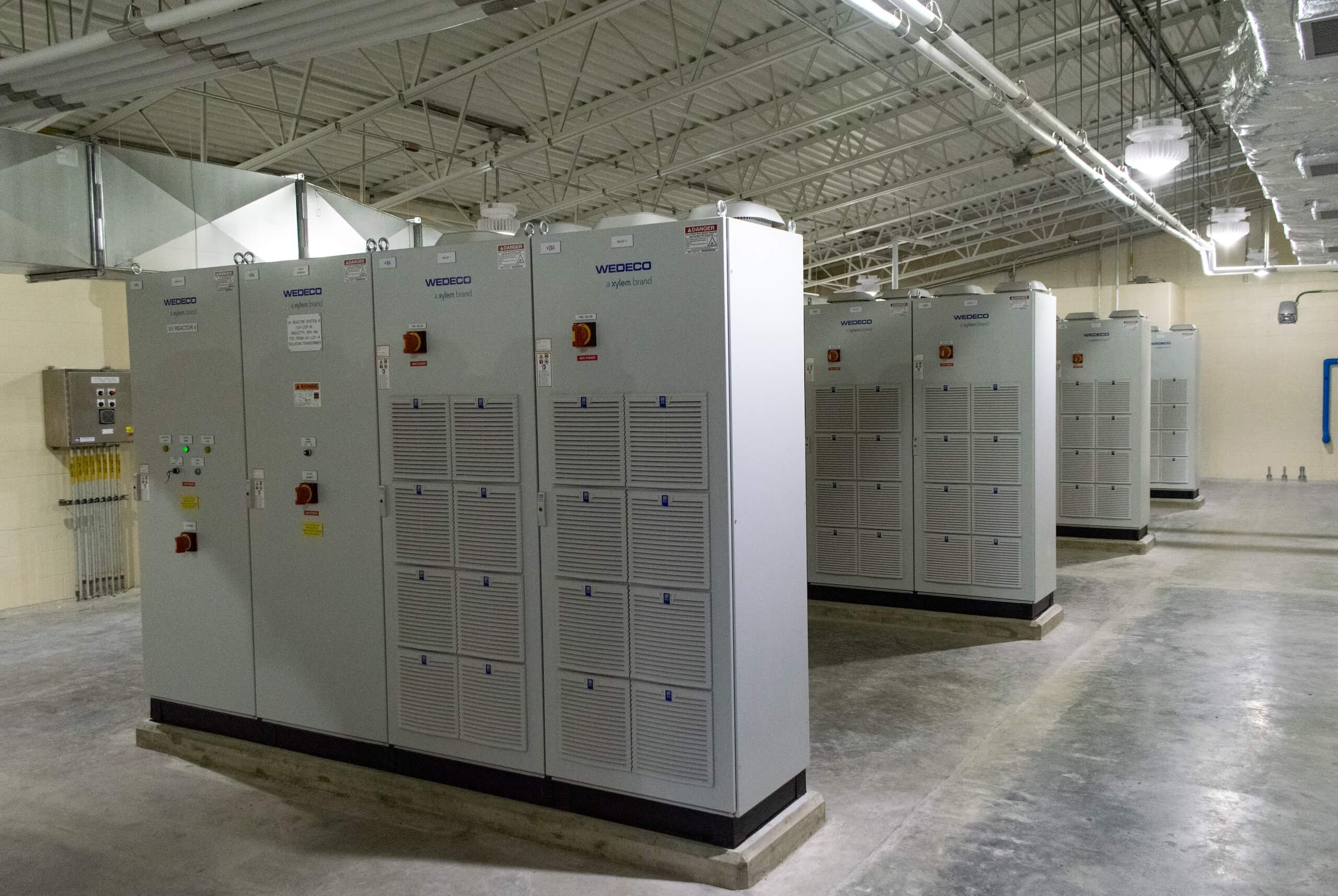 Large industrial electrical control panels labeled Wedeco are lined up in a clean, spacious room with concrete floors, beige walls, and exposed ceiling pipes.