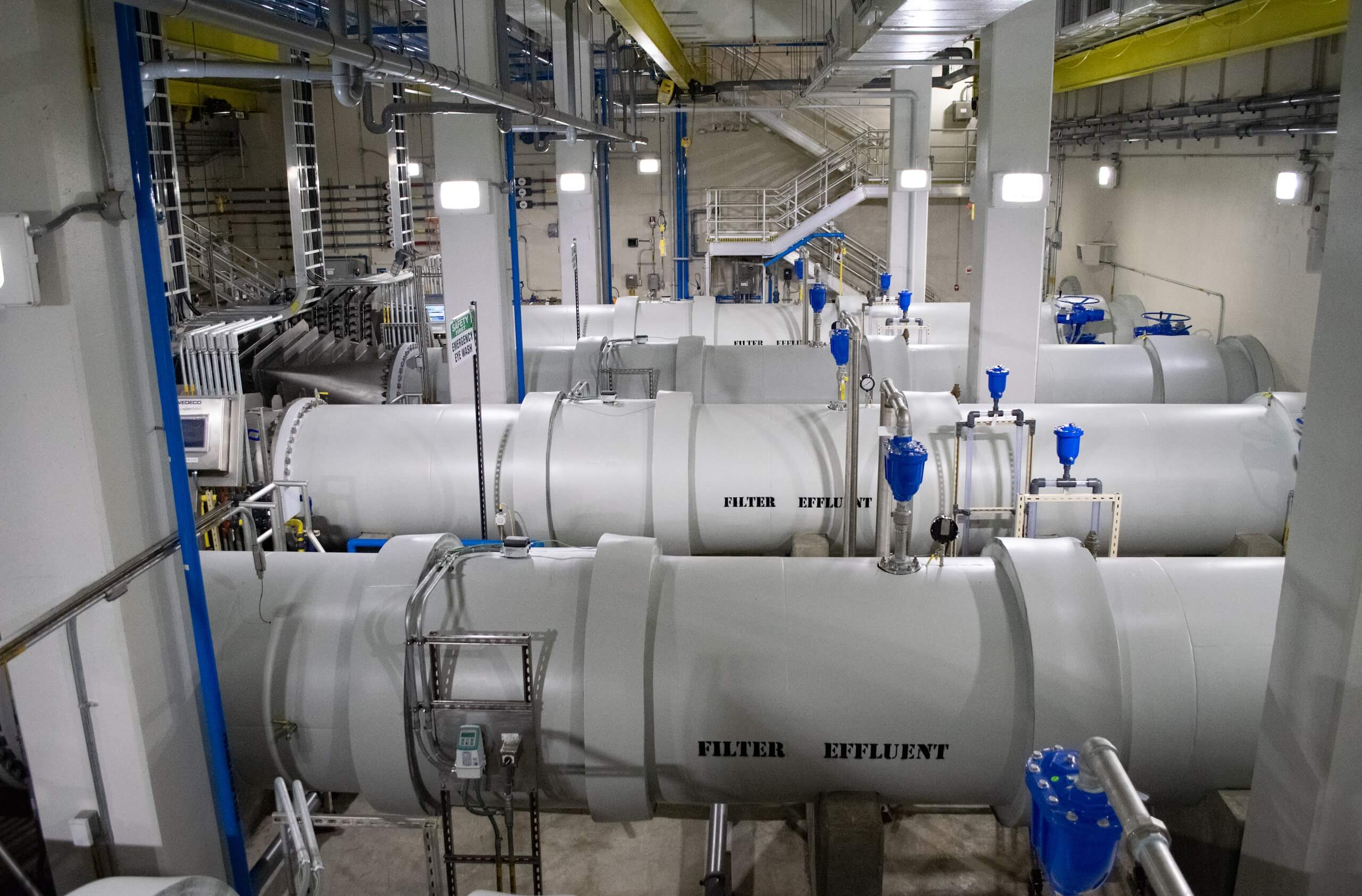 Large industrial facility with white cylindrical tanks labeled FILTER and EFFLUENT, blue valves, pipes, metal walkways, and bright overhead lighting. Stairs and platforms are visible in the background.