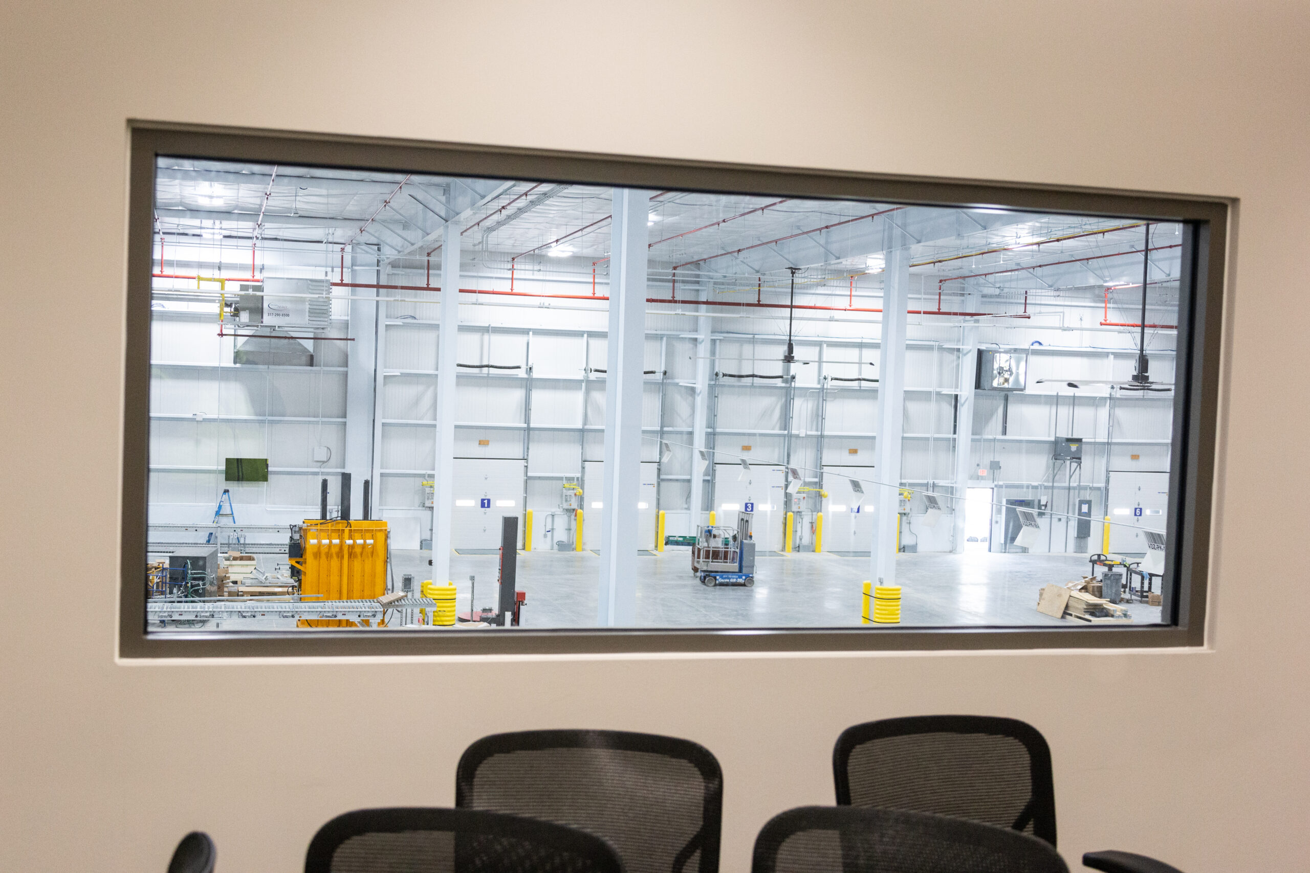 A large, empty industrial warehouse is viewed through a window from a conference room with black office chairs. The warehouse has high ceilings, white walls, and minimal equipment scattered throughout.