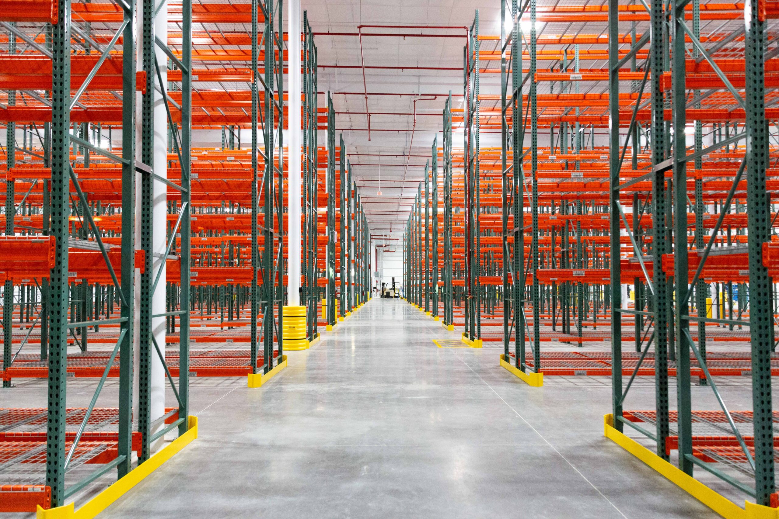 Wide view of an empty warehouse with tall rows of orange and green metal shelving units, bright lighting, and a polished concrete floor. The shelves are unused, and the space appears clean and organized.
