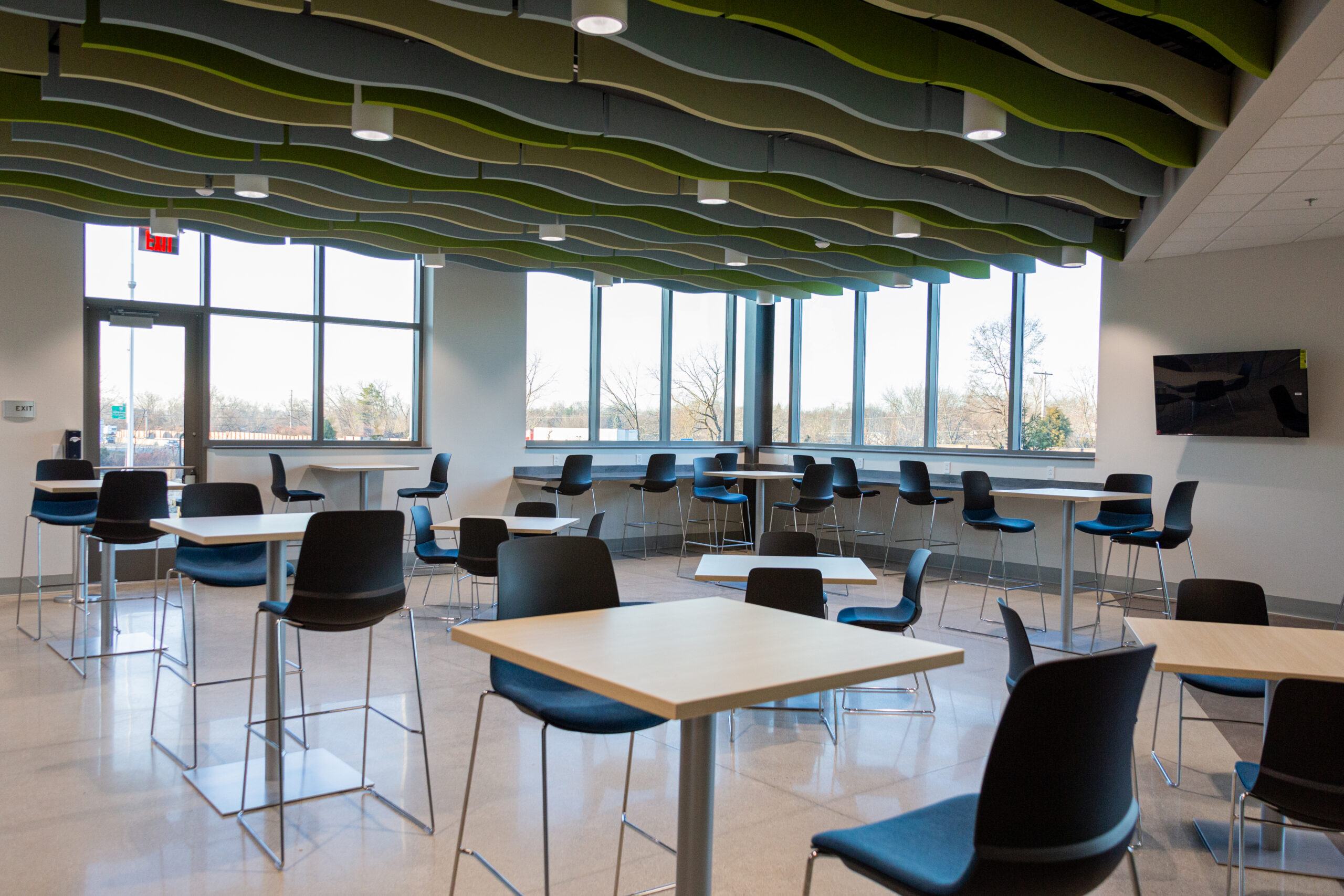 Modern cafeteria with square tables and black chairs, large windows letting in natural light, and a green wavy ceiling design. A wall-mounted TV is on the right.