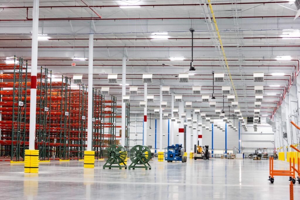 A spacious, well-lit warehouse interior with empty orange storage racks on the left and various equipment and machinery scattered across the clean, polished floor.