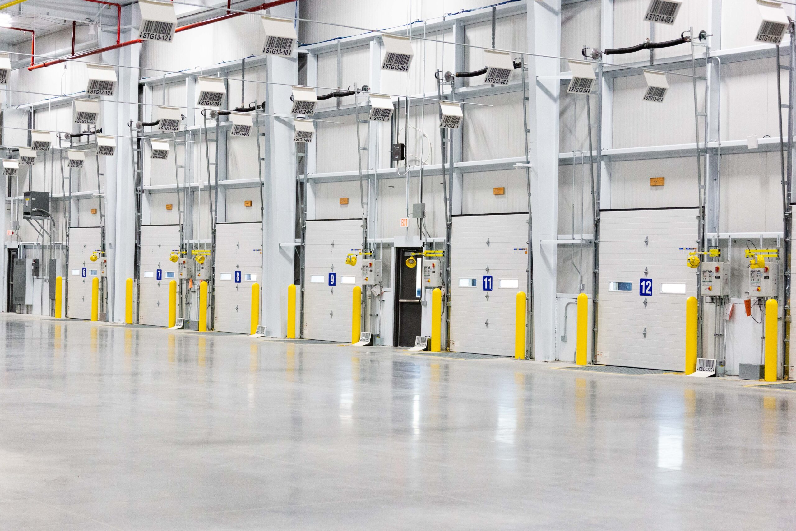 Interior of a large, empty warehouse with numbered loading dock doors, yellow safety barriers, and polished concrete floors.
