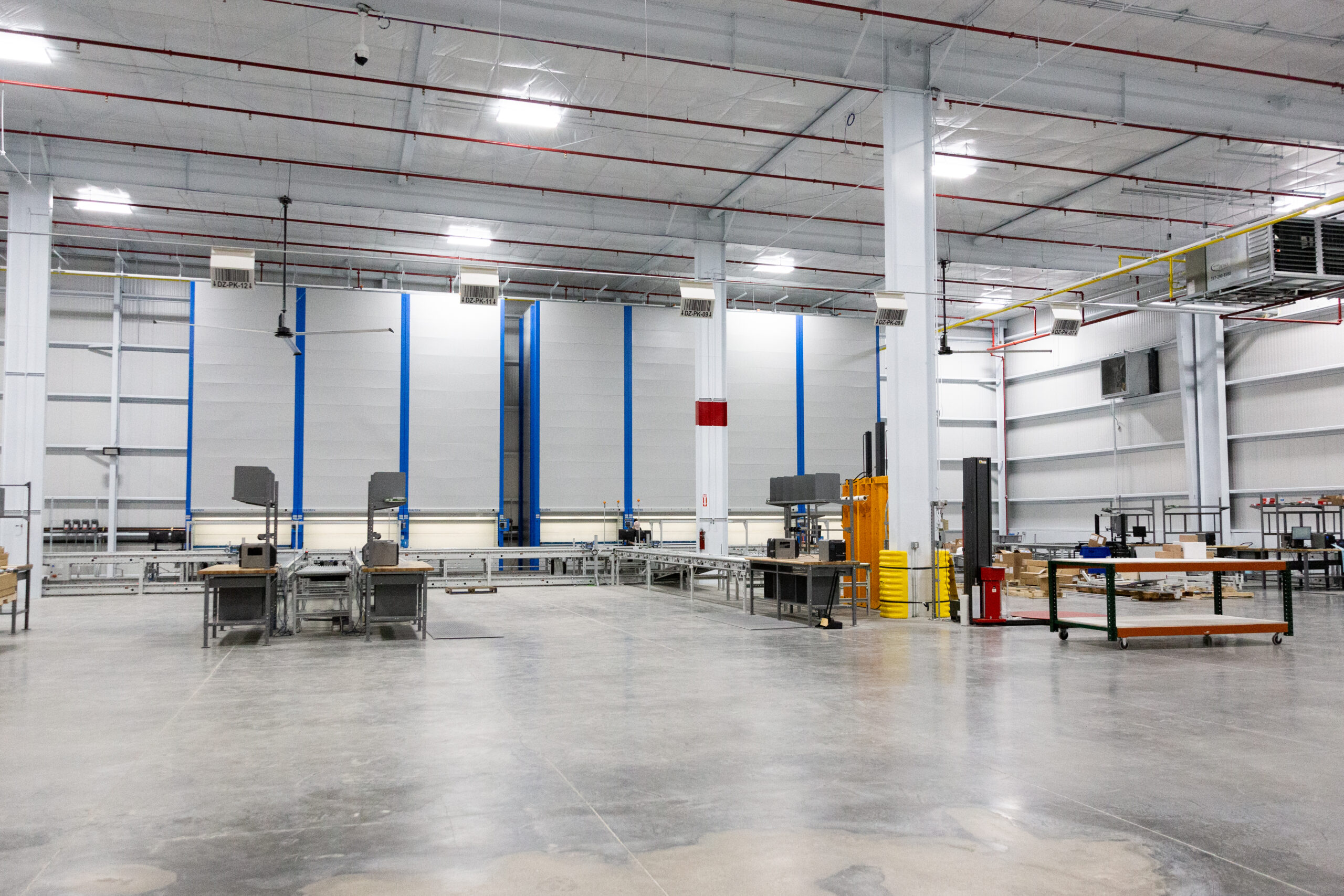 A spacious, modern industrial warehouse with empty workstations, metal tables, carts, machinery, and concrete floors under bright overhead lighting.