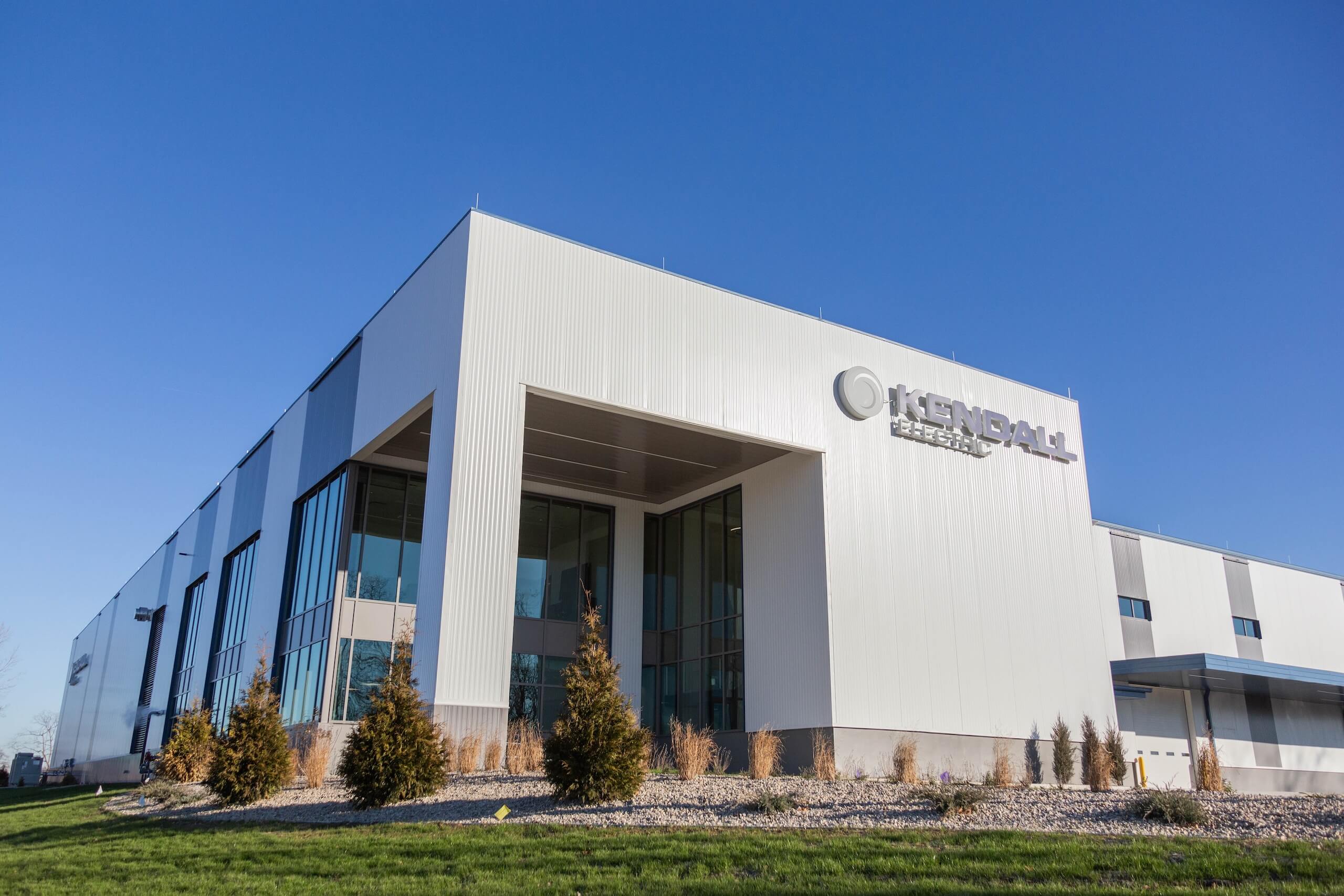 Modern white industrial building with large windows, labeled Kendall Electric, sits under a clear blue sky, surrounded by green grass, shrubs, and small trees.