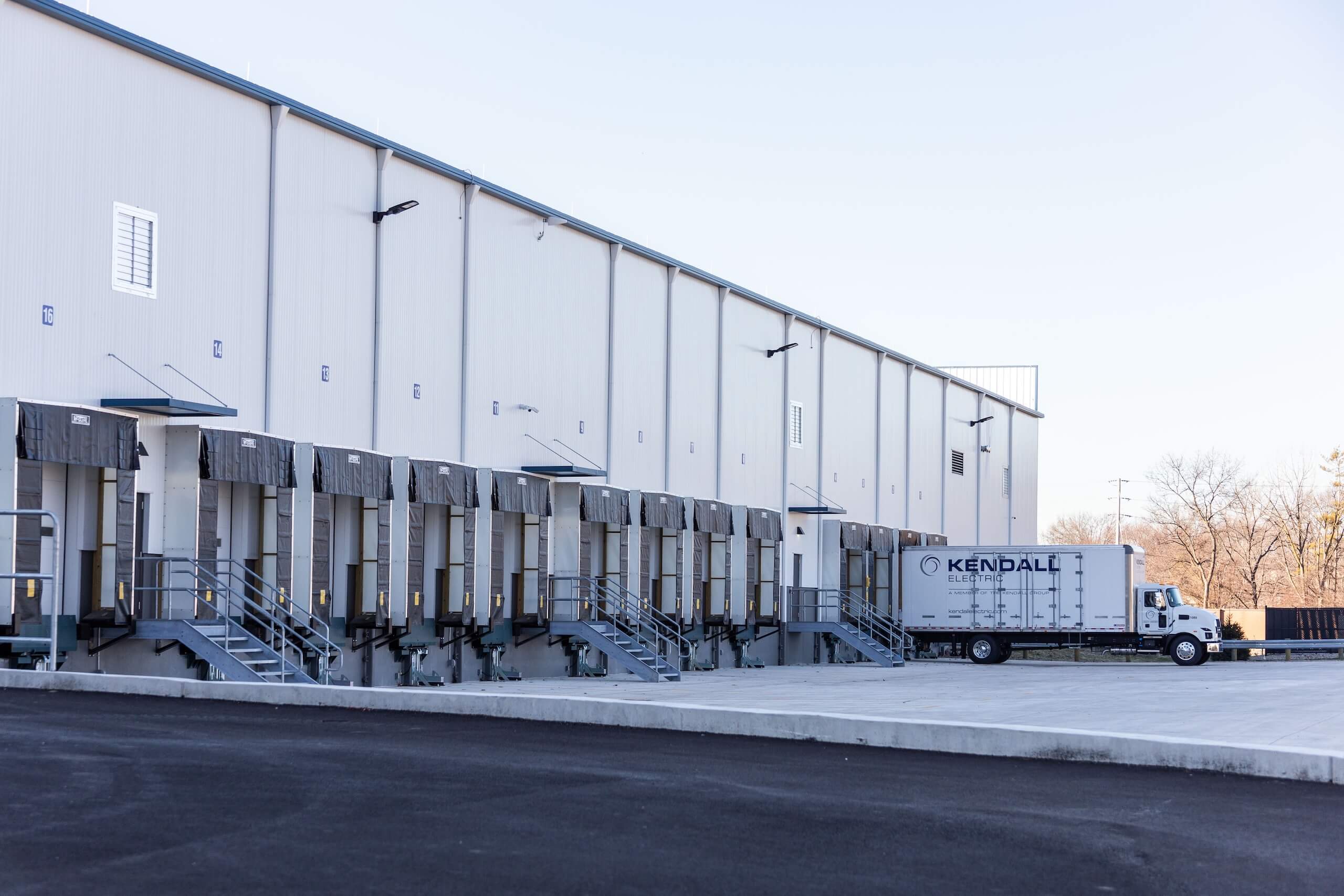 A white truck labeled Kendall is parked at a loading dock outside a large industrial warehouse with multiple loading bays and metal stairs.