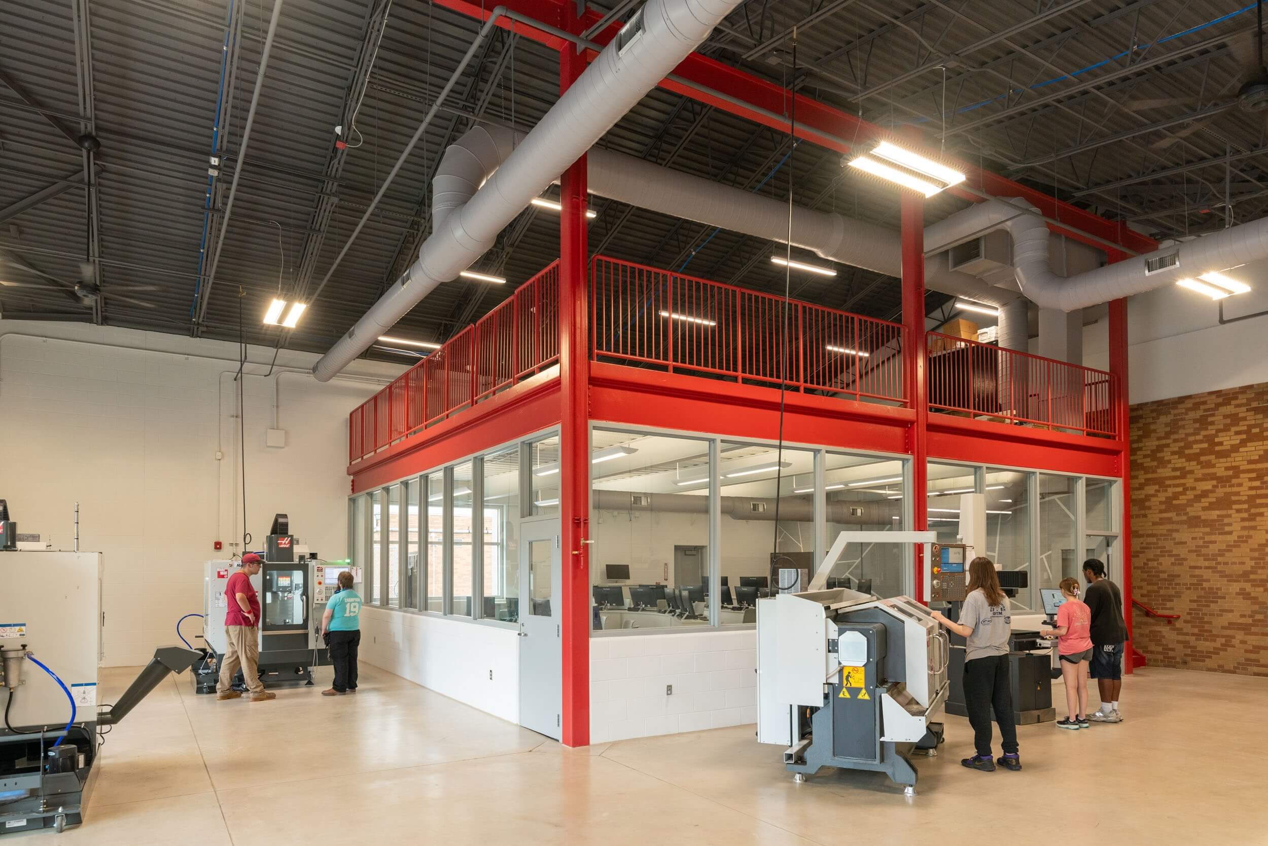 People work with machines in a spacious industrial room with red railings, glass-walled office space, and exposed ductwork on the ceiling. Some stand near equipment, while others observe through the office windows.