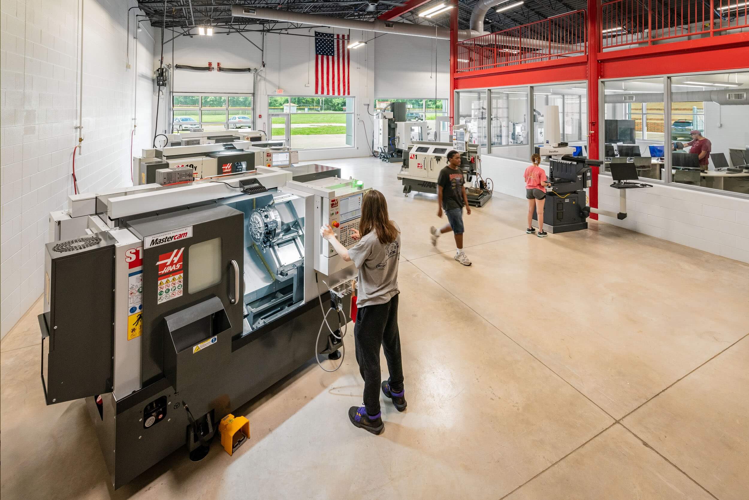 Several students operate machines in a spacious, well-lit workshop with large windows, white walls, and an American flag hanging. A few people work at desks behind glass in a separate office area.