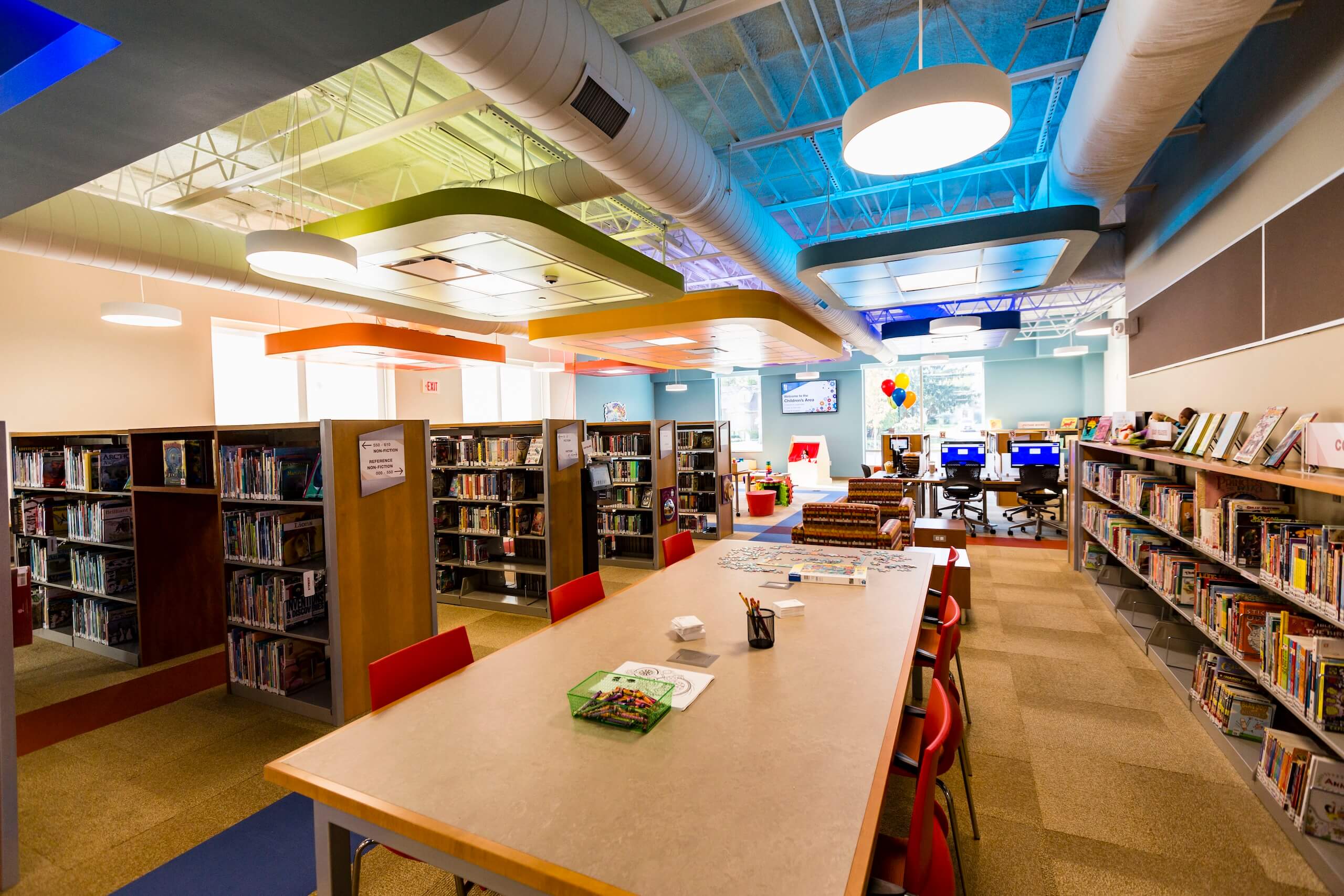 Colorful modern library with bookshelves, a central table with chairs and art supplies, computers in the back, and large windows letting in natural light.