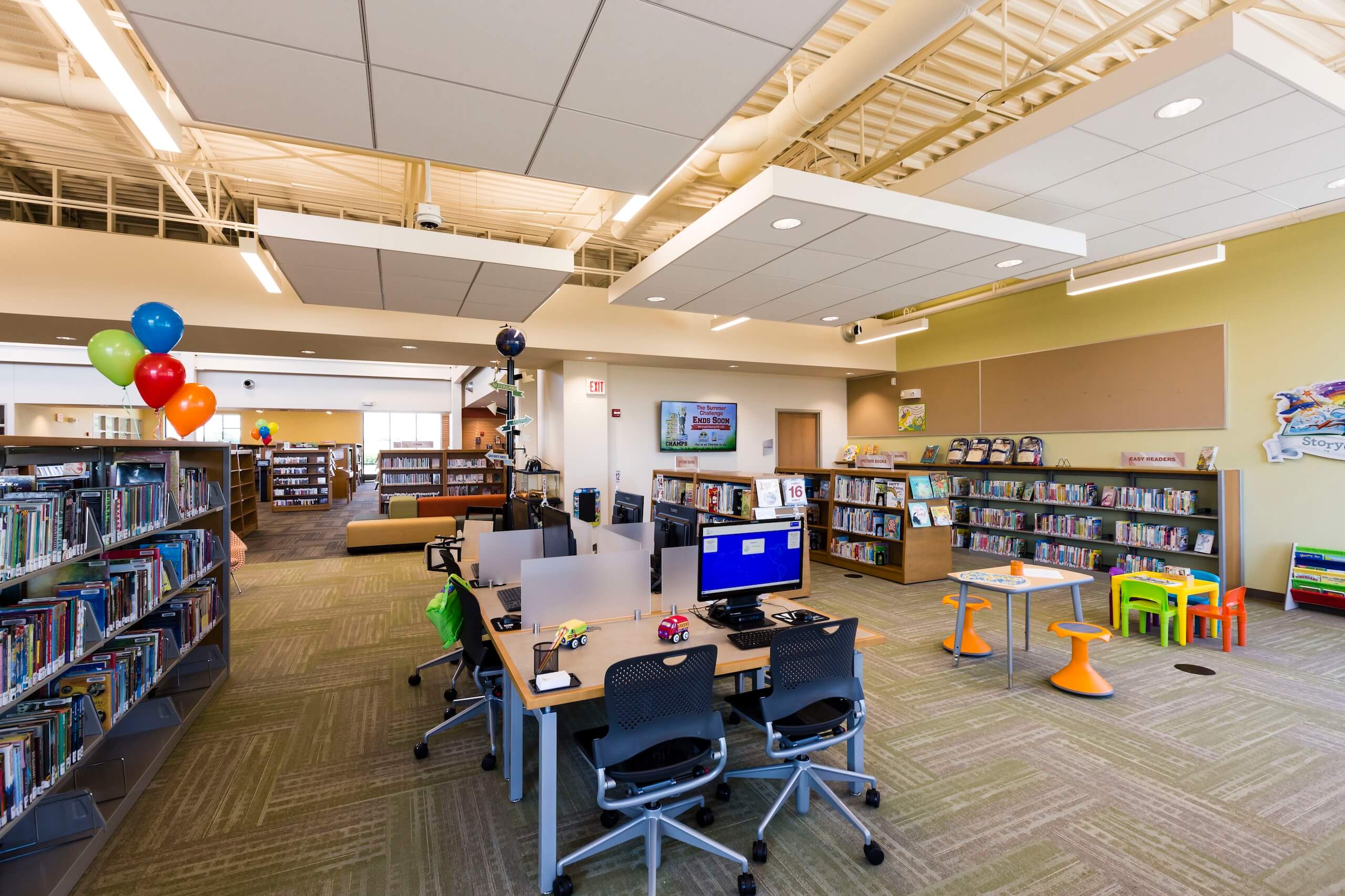 A modern library childrens area with bookshelves, colorful balloons, computers on desks, and a small play table with chairs. Bright lighting and organized spaces create a welcoming atmosphere.