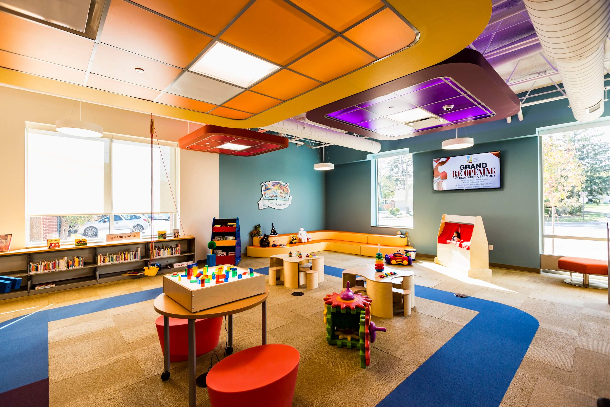 Bright, colorful childrens playroom with toys, bookshelves, play tables, a TV displaying Grand Re-Opening, and large windows letting in natural light. Walls and ceiling are decorated in vibrant patterns.