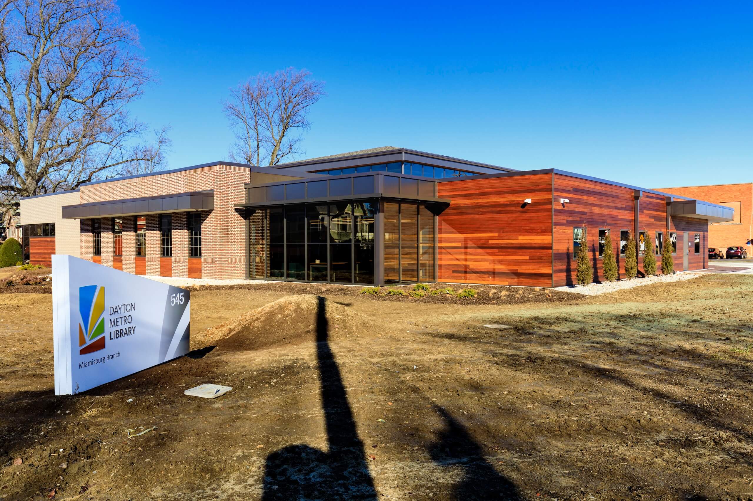 Modern library building with large windows and wood paneling, under a clear blue sky. A sign in front reads Dayton Metro Library, Miamisburg Branch with landscaping and bare trees visible.