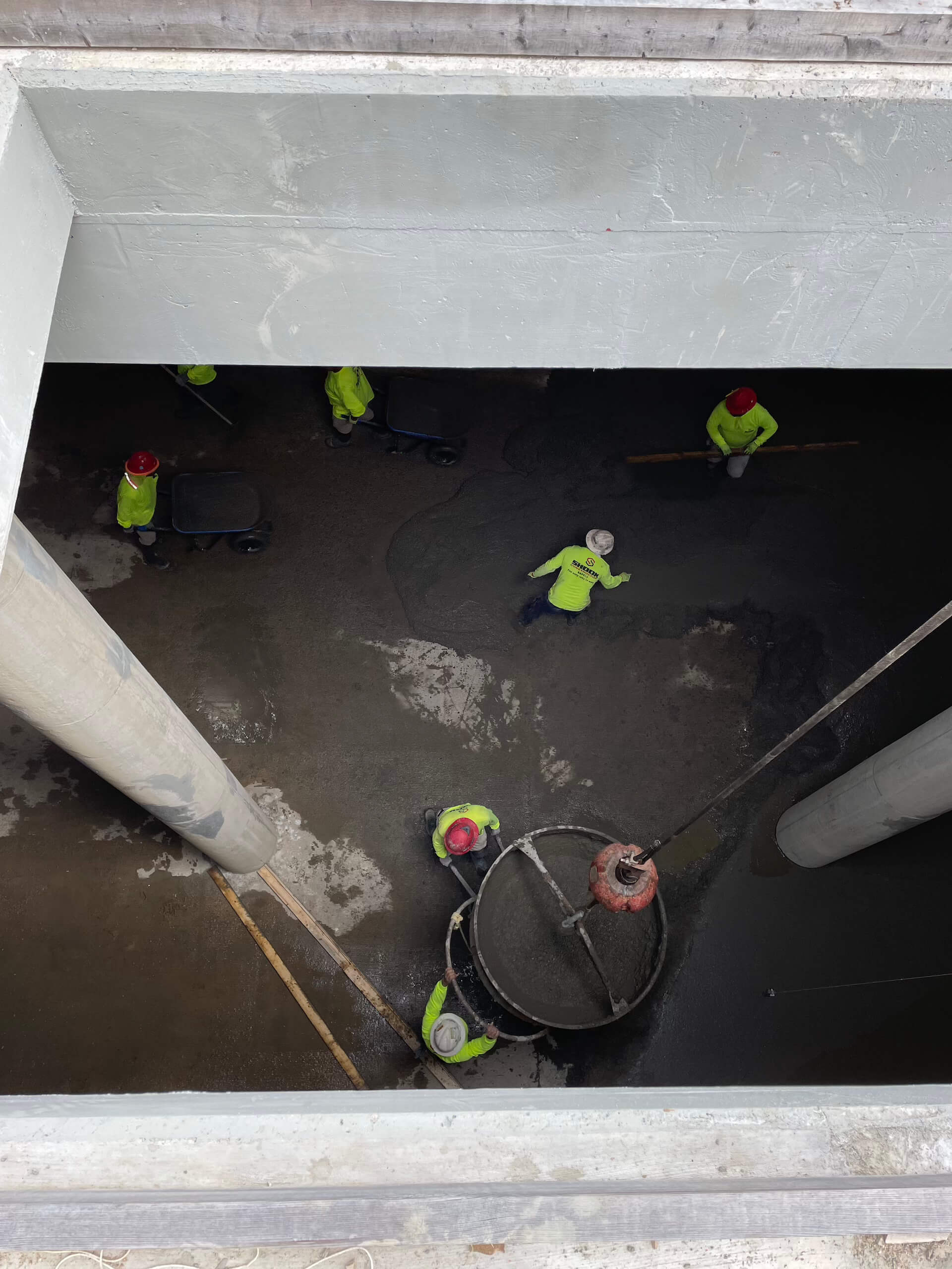 Five construction workers in bright green safety gear and helmets are seen from above, working with concrete inside a large, unfinished structure with two big columns. A suspended concrete bucket is being used.