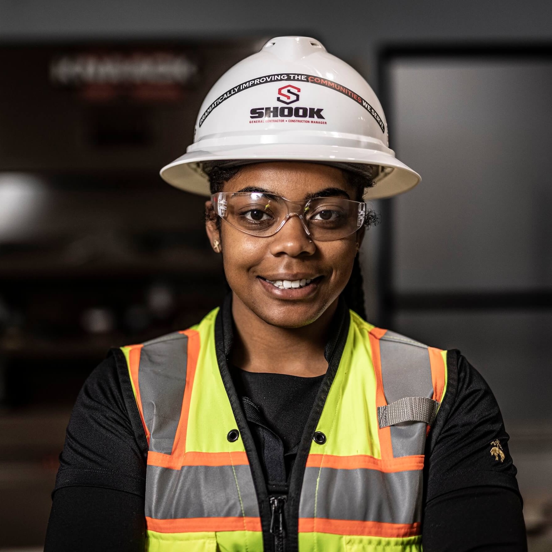 A woman wearing a hard hat, safety glasses, and a reflective vest smiles at the camera in an indoor construction setting.