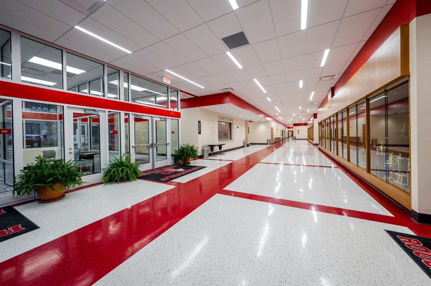 A wide, brightly lit school hallway with red and white flooring, trophy cases on the right, glass-walled rooms on the left, and a few green potted plants along the corridor.