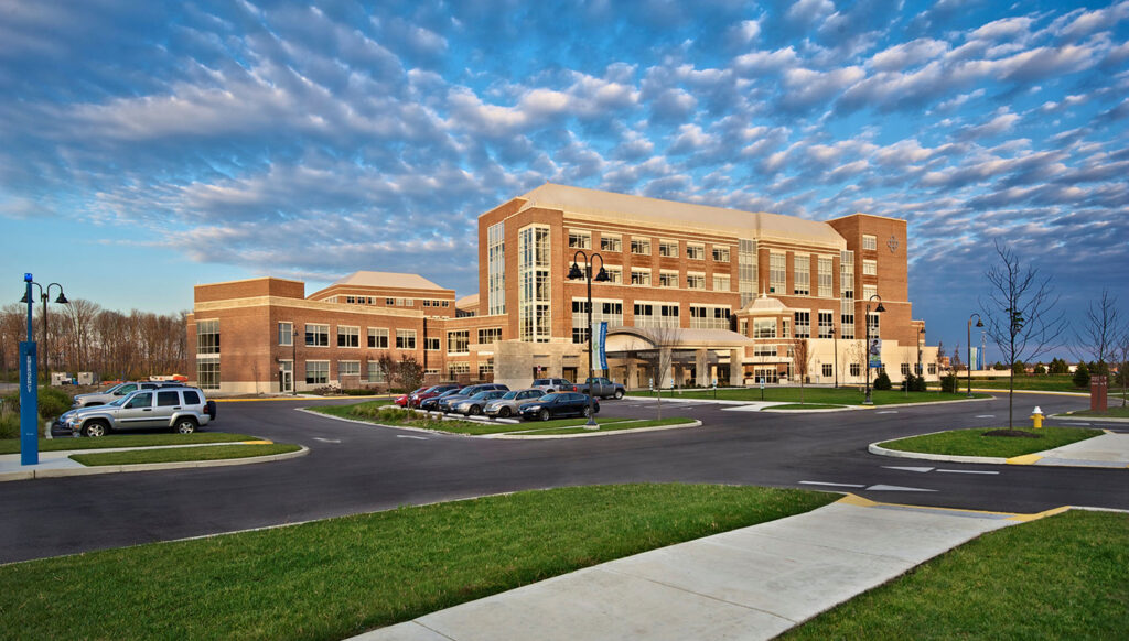 A modern, multi-story brick building with large windows, a main entrance, and several parked cars in front, set against a blue sky with scattered clouds. Green lawns and sidewalks surround the structure.