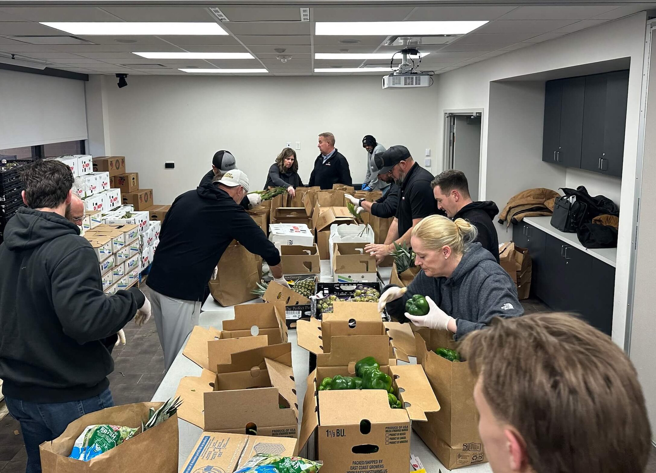 A group of people pack fresh produce and groceries into paper bags at a long table in a well-lit room, working together as part of a food distribution or volunteer event.