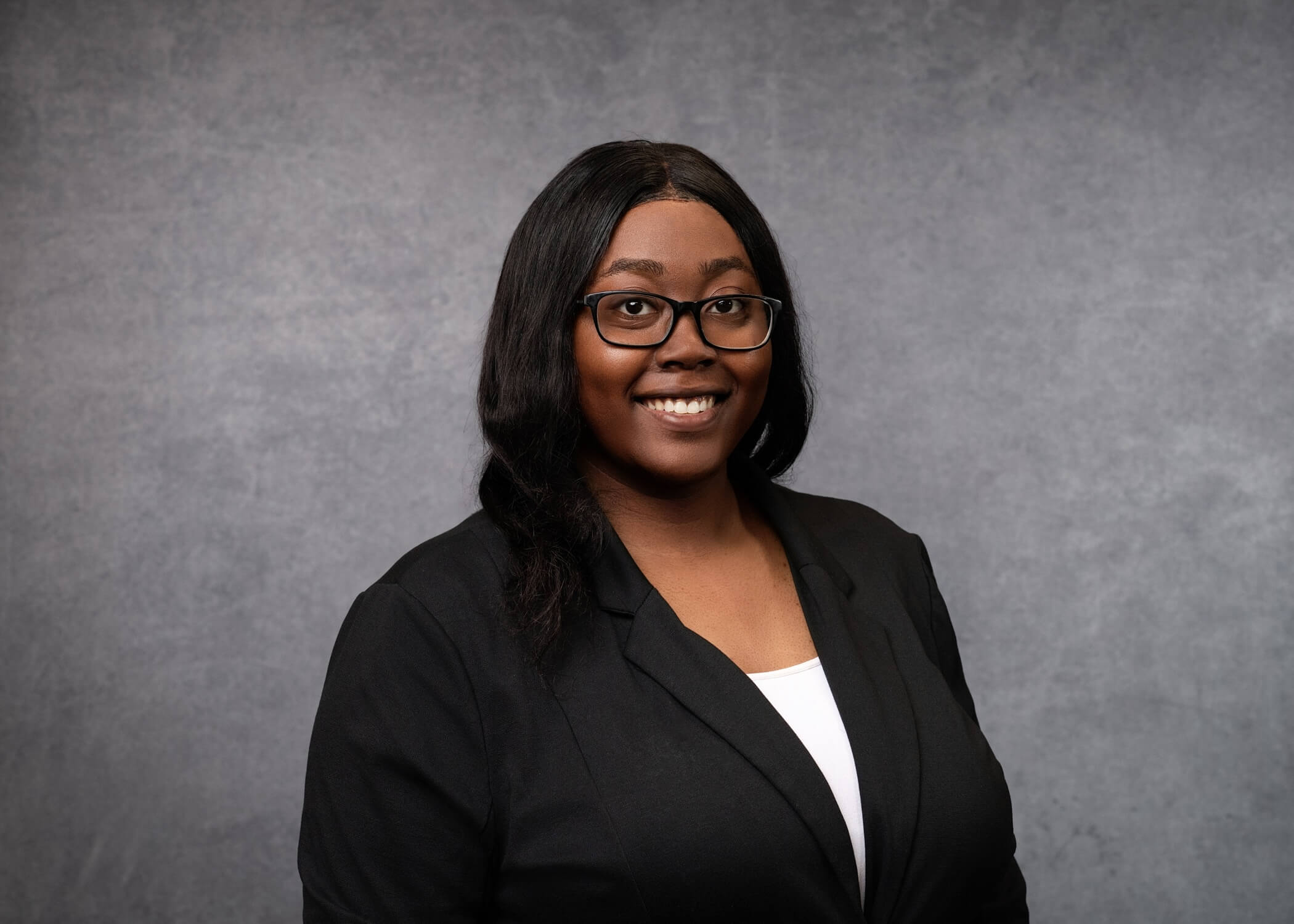 A woman wearing glasses, a black blazer, and a white top smiles in front of a plain gray background.