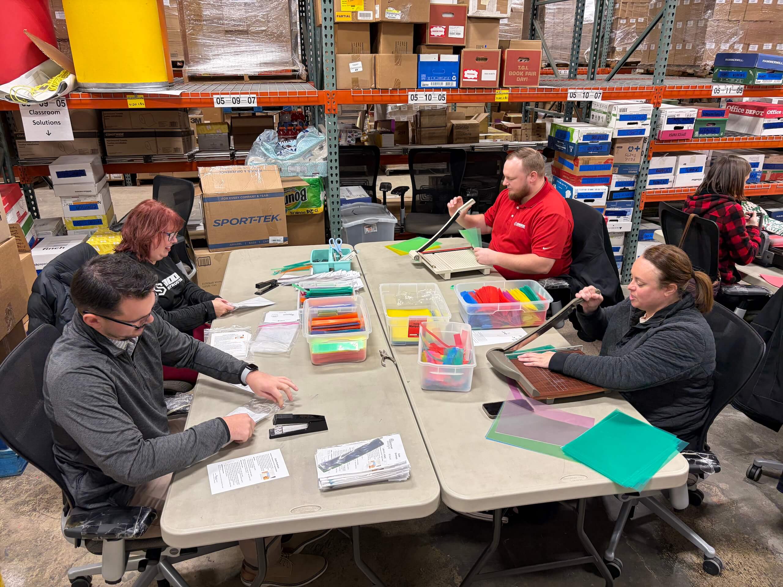 Four people sit at tables in a warehouse, assembling folders and paperwork with colorful plastic bins in front of them. Shelves filled with boxes and supplies are visible in the background.