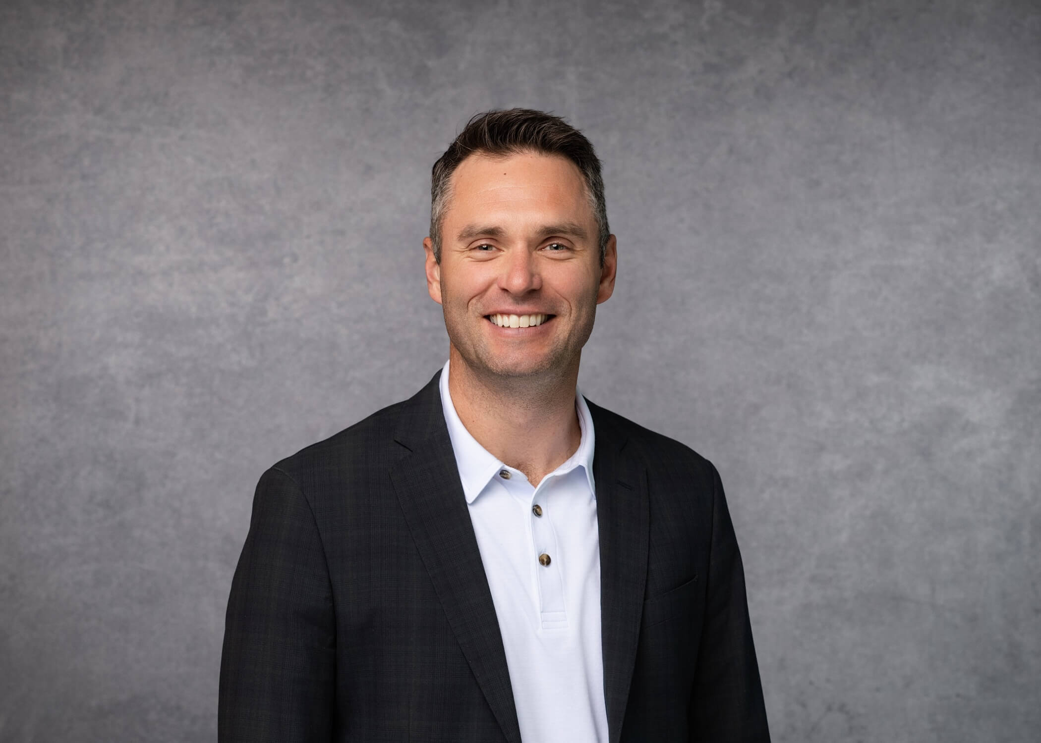 A man with short brown hair smiles at the camera, wearing a dark blazer over a white collared shirt. The background is a plain gray studio backdrop.