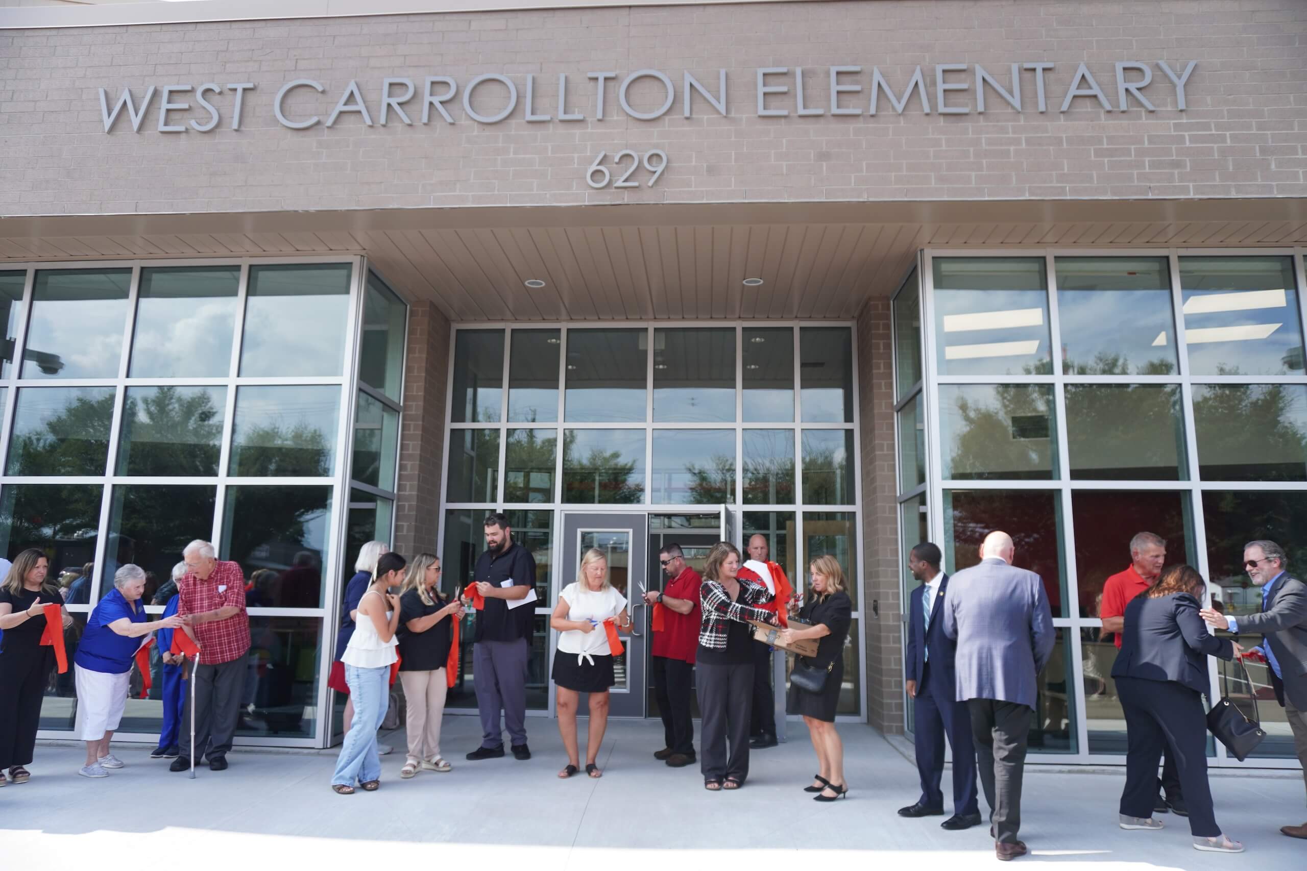 A group of people gather outside the entrance of West Carrollton Elementary, standing and talking in front of the buildings glass doors and windows.