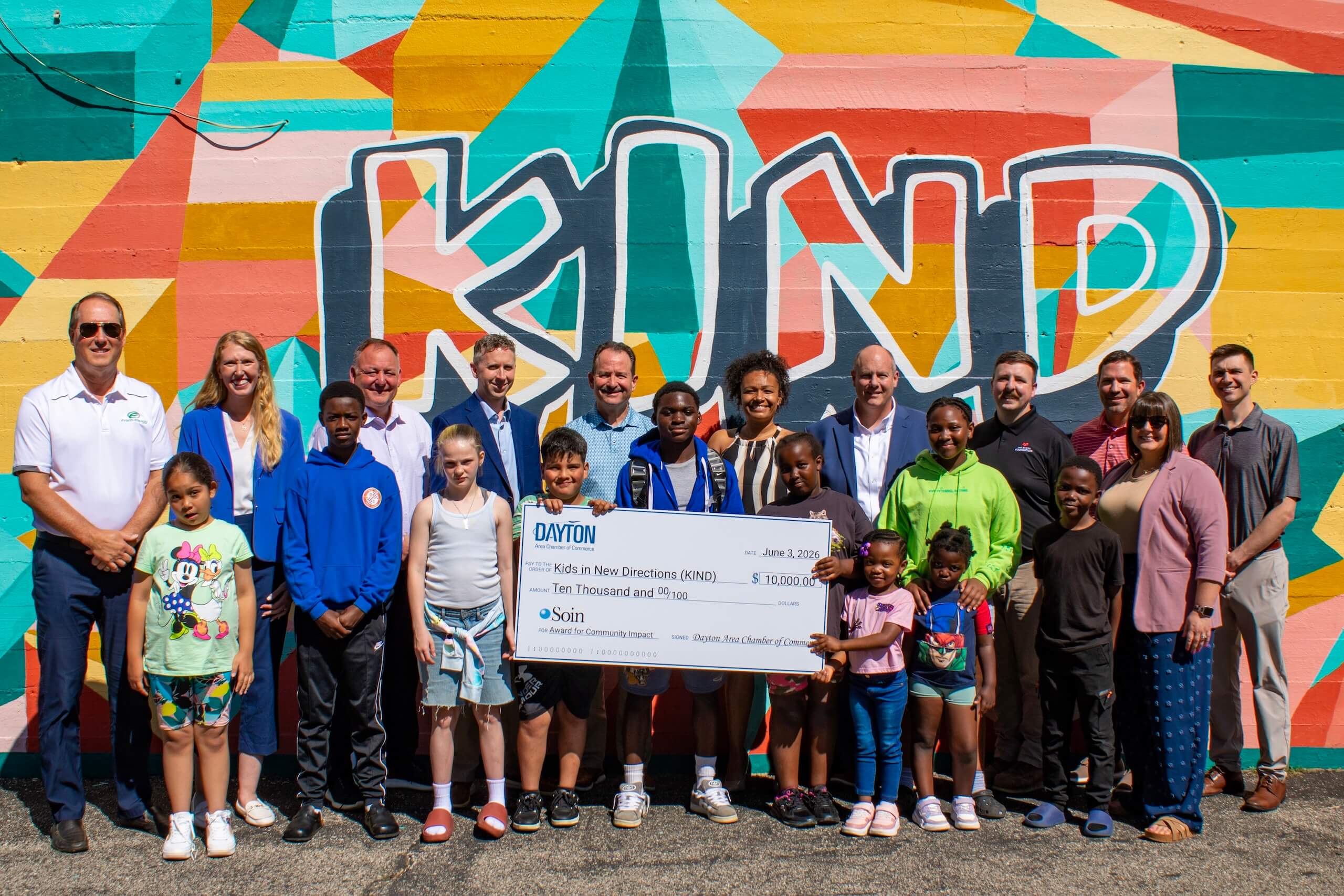 A group of adults and children pose outdoors in front of a colorful mural that says KIND, holding a large ceremonial check for $10,000 from SKIN and Dayton organizations.