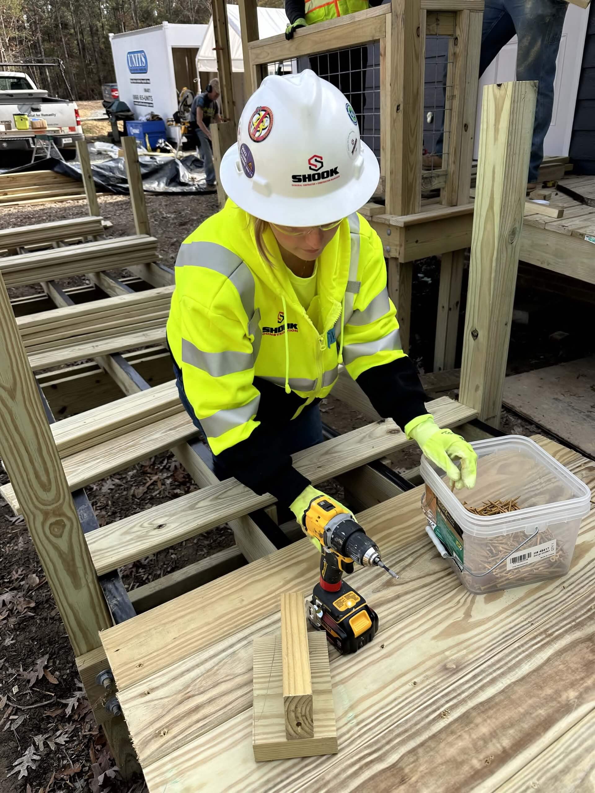 A construction worker in a neon safety jacket and hard hat prepares to drill wood on an outdoor staircase, with screws in a plastic container nearby.
