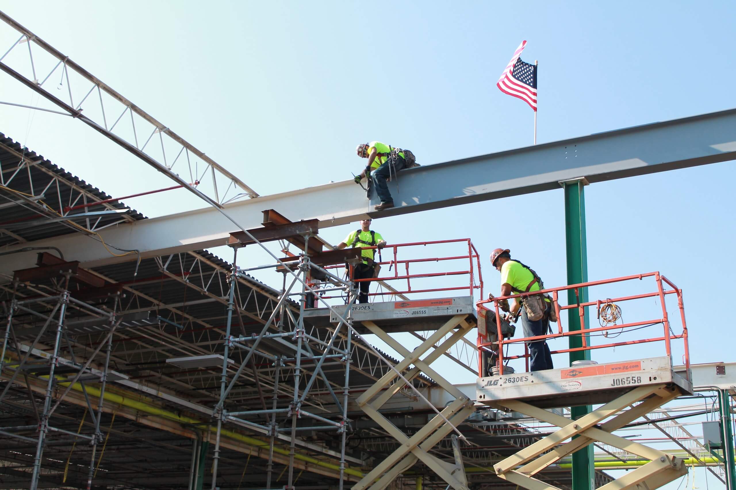 Three construction workers in safety gear work on a steel beam atop scaffolding and lifts, with an American flag flying above the structure against a clear blue sky.