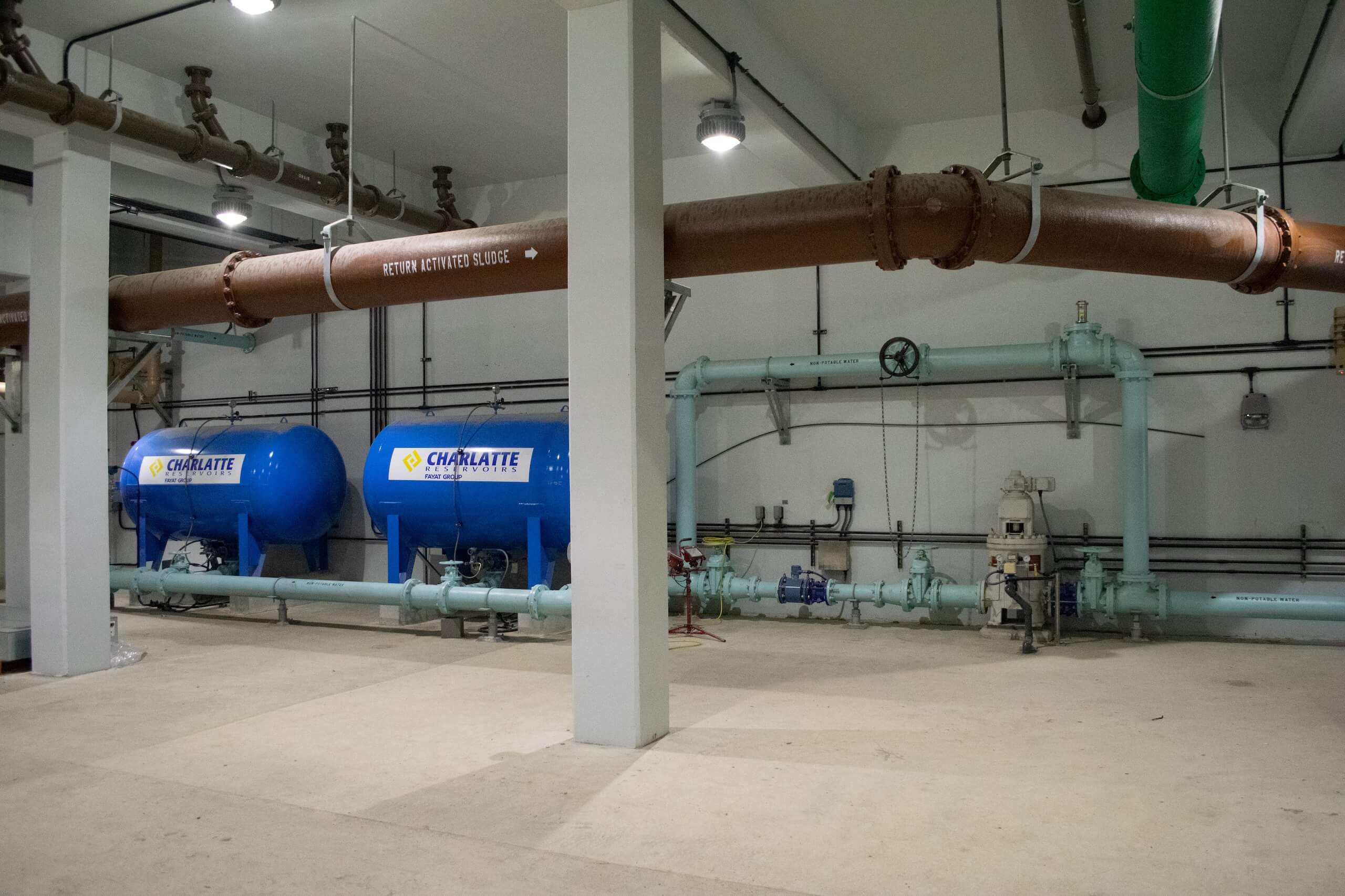 Large industrial room with tanks labeled Chalklate, brown and green pipes overhead, and light blue machinery and pipes along the back wall on a concrete floor under bright overhead lights.