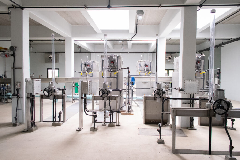 Industrial facility interior with white walls, large pipes, valves, metal machinery, and equipment, under natural light from ceiling openings. The area appears clean and organized.