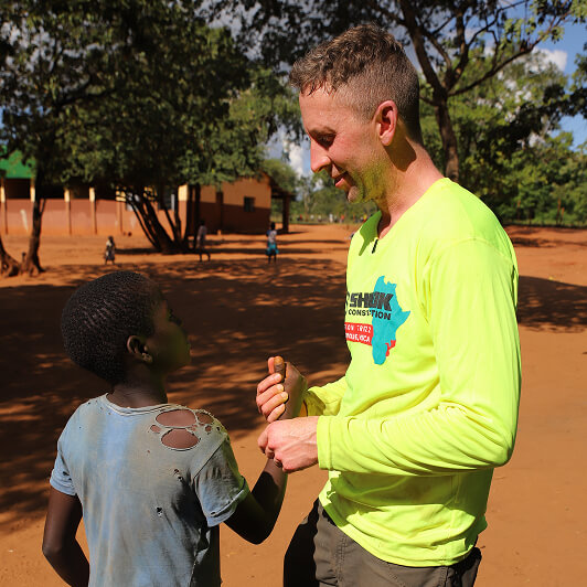 A man in a bright yellow shirt talks to a child in a worn blue shirt outside on a sunny day, with trees and buildings in the background.