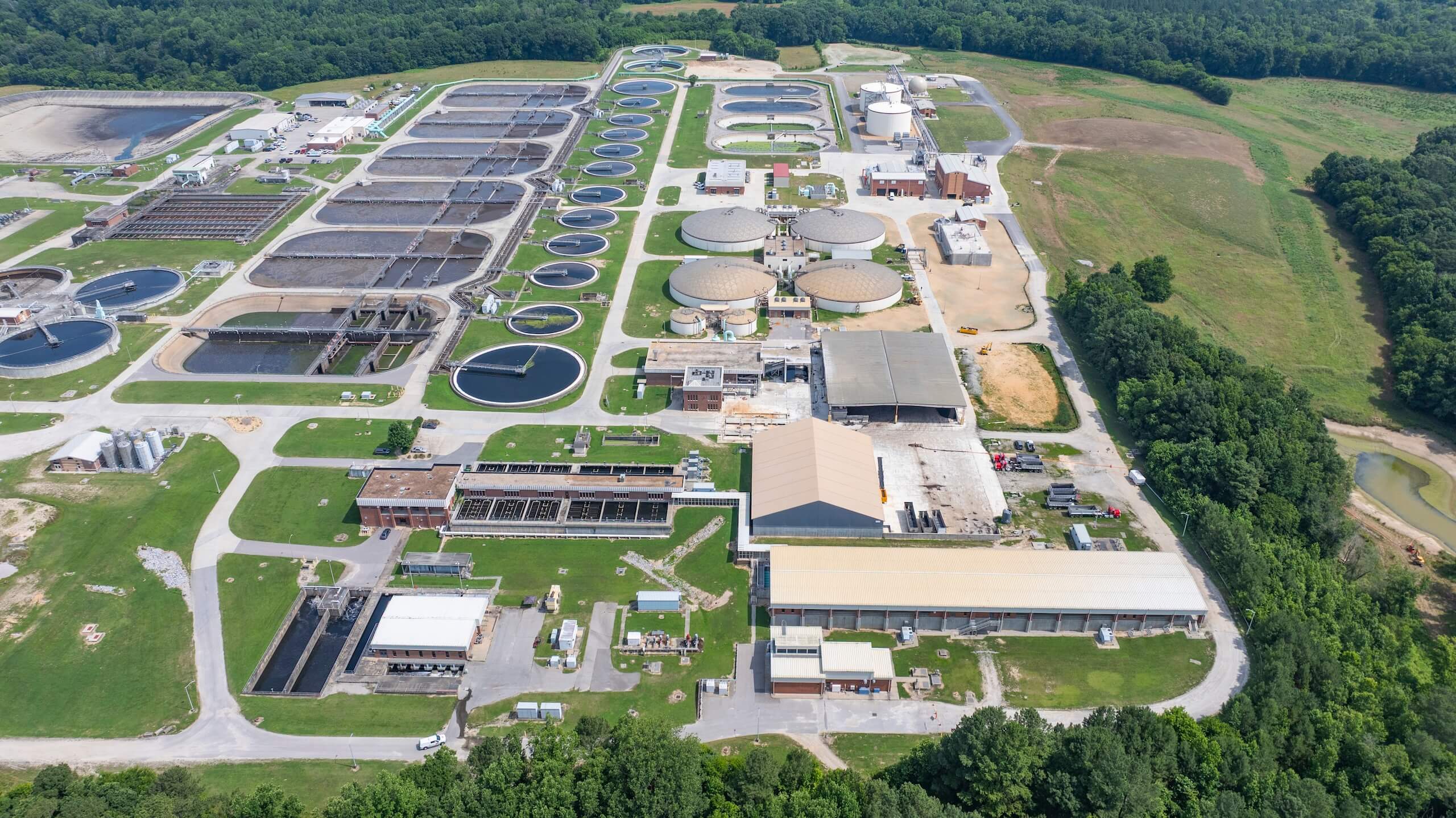 Aerial view of a large wastewater treatment plant with multiple circular and rectangular tanks, surrounding buildings, and green areas, bordered by trees and grassy fields.